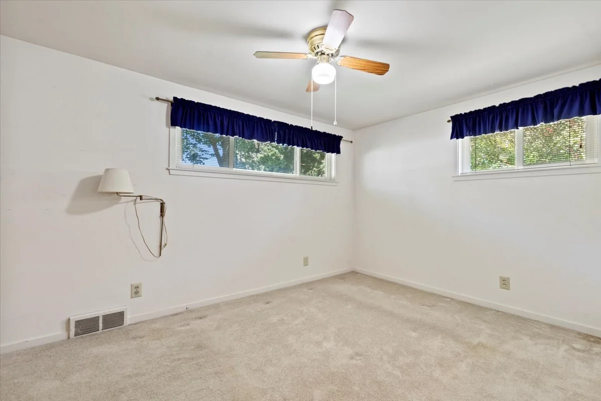 Empty bedroom with white walls, beige carpet, two windows with blue curtains, ceiling fan with light, wall-mounted lamp, and a vent near the floor.