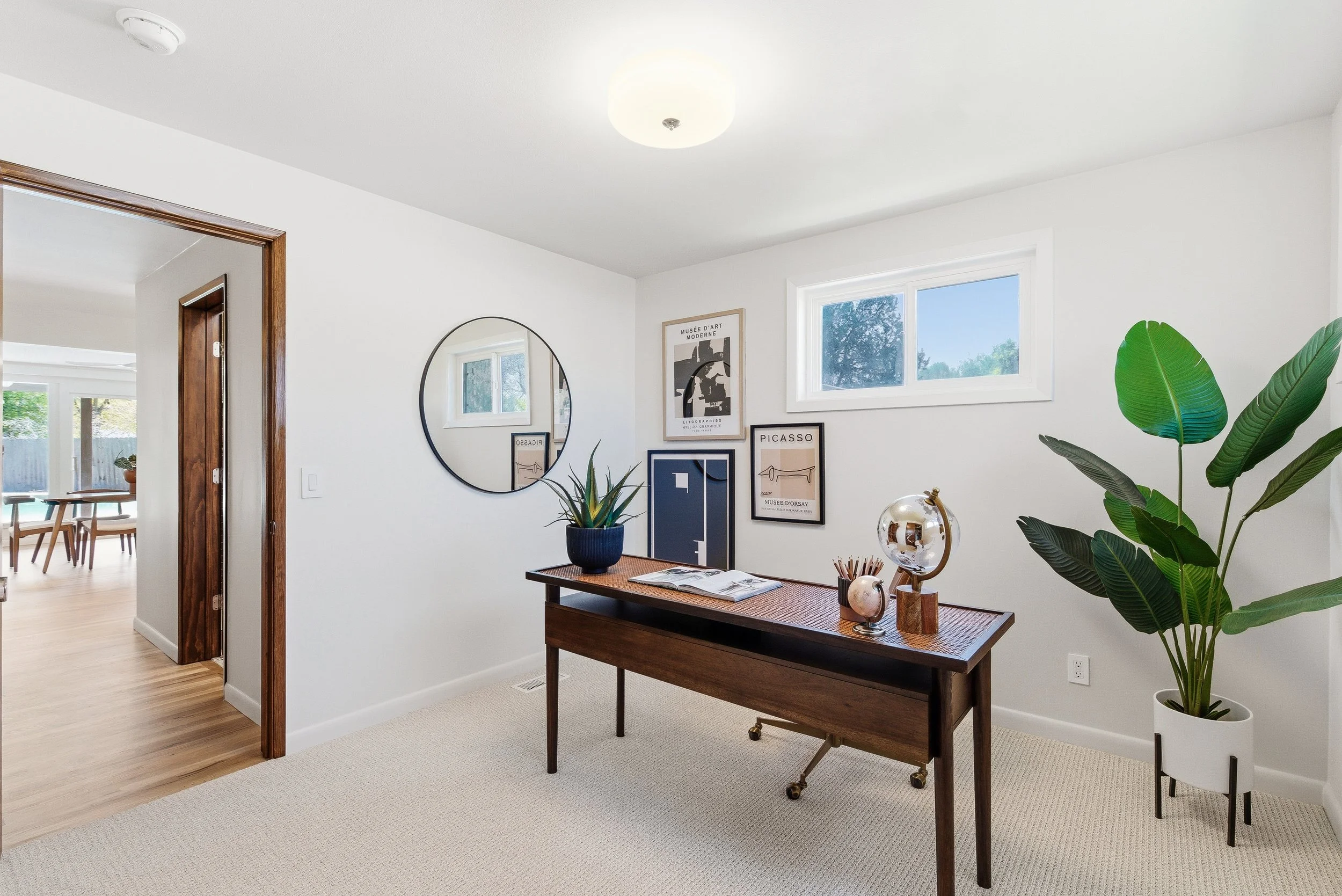 A well-lit interior room with white walls, a wooden table, a large green potted plant, framed art on the wall, a mirror, and large windows allowing natural light.
