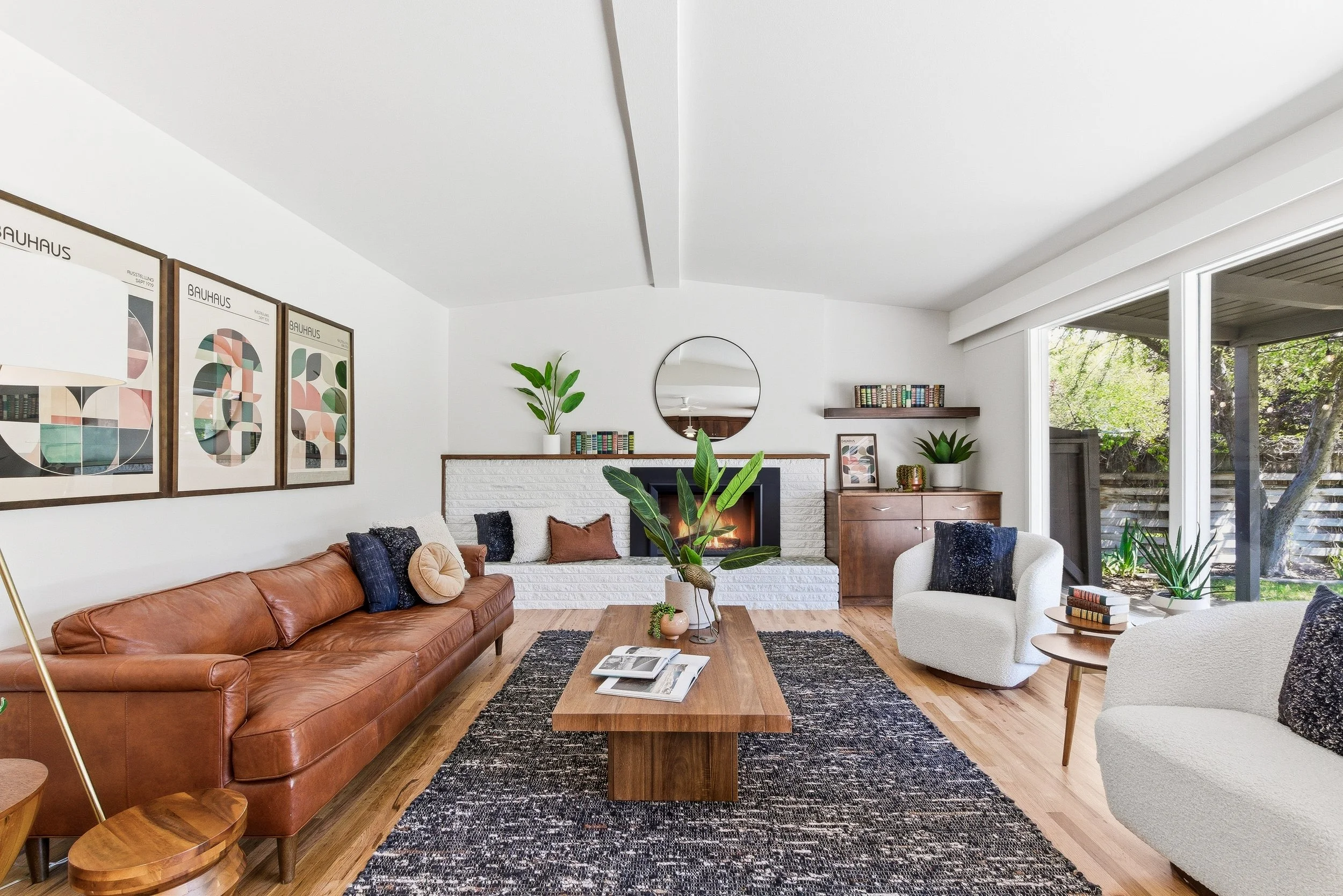 Living room with white walls, large windows showing trees, wooden floor, brown leather sofa, white armchairs, wooden coffee table, plants, framed artwork, and mirror above the fireplace.