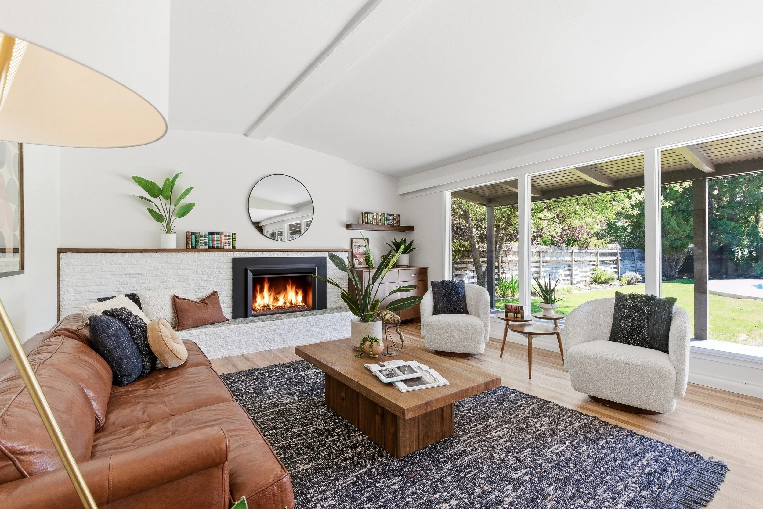 Living room with a leather sofa, two white armchairs, a wooden coffee table, a fireplace, bookshelves, and large windows showing a backyard with trees and a fence.