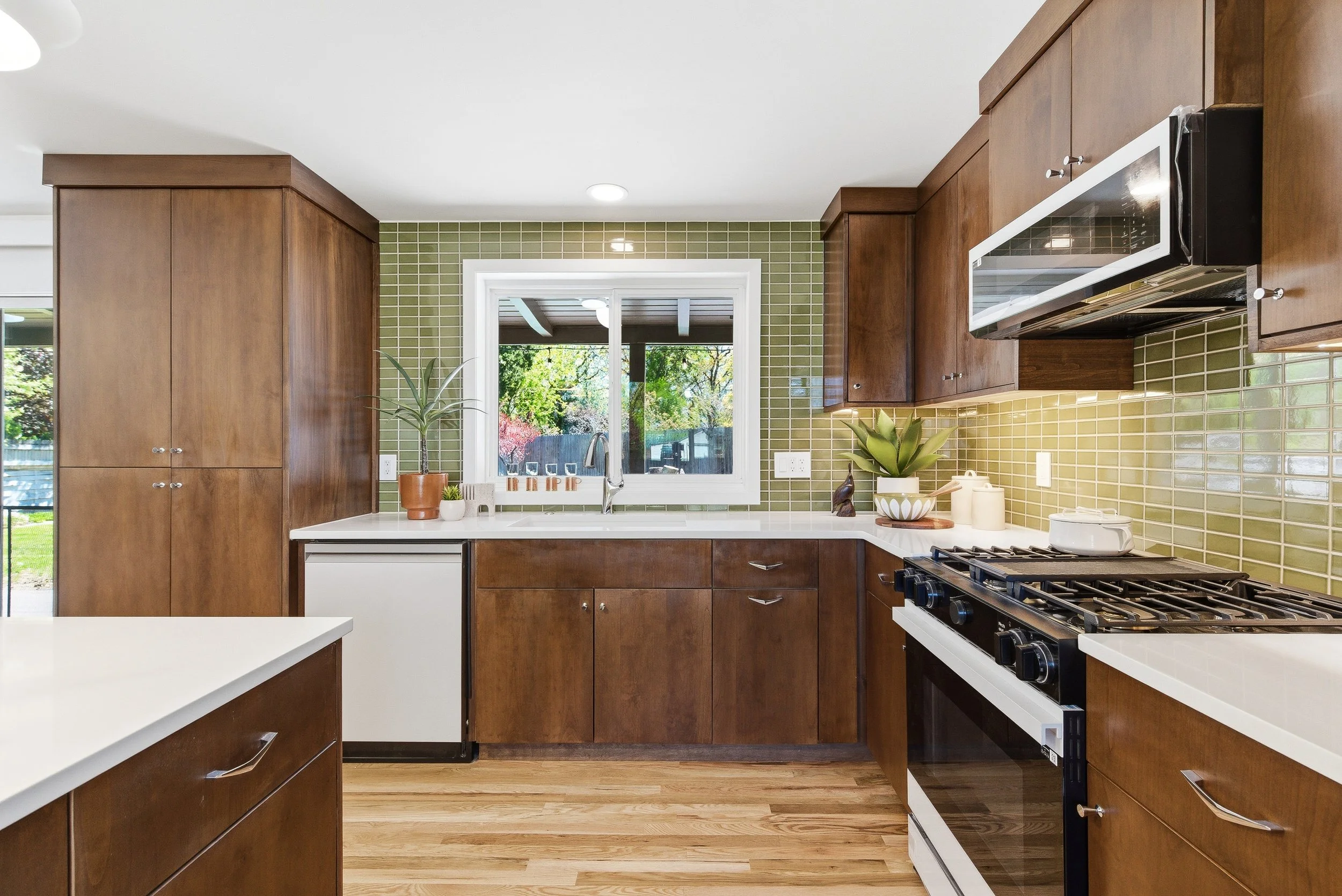 Modern kitchen with wooden cabinets, white countertops, green tile backsplash, and a window overlooking a backyard with trees.