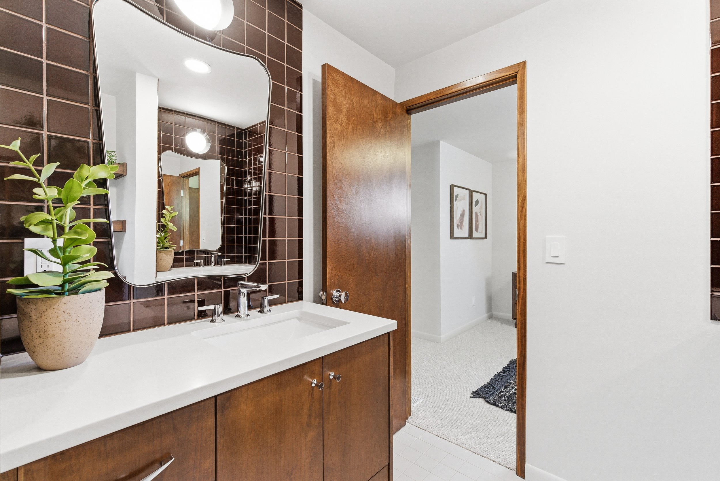 A bathroom vanity with a white countertop, a brown tiled wall, and a mirror. There is a potted plant on the countertop, a wooden door partly open, and two framed pictures on the wall in an adjacent room.
