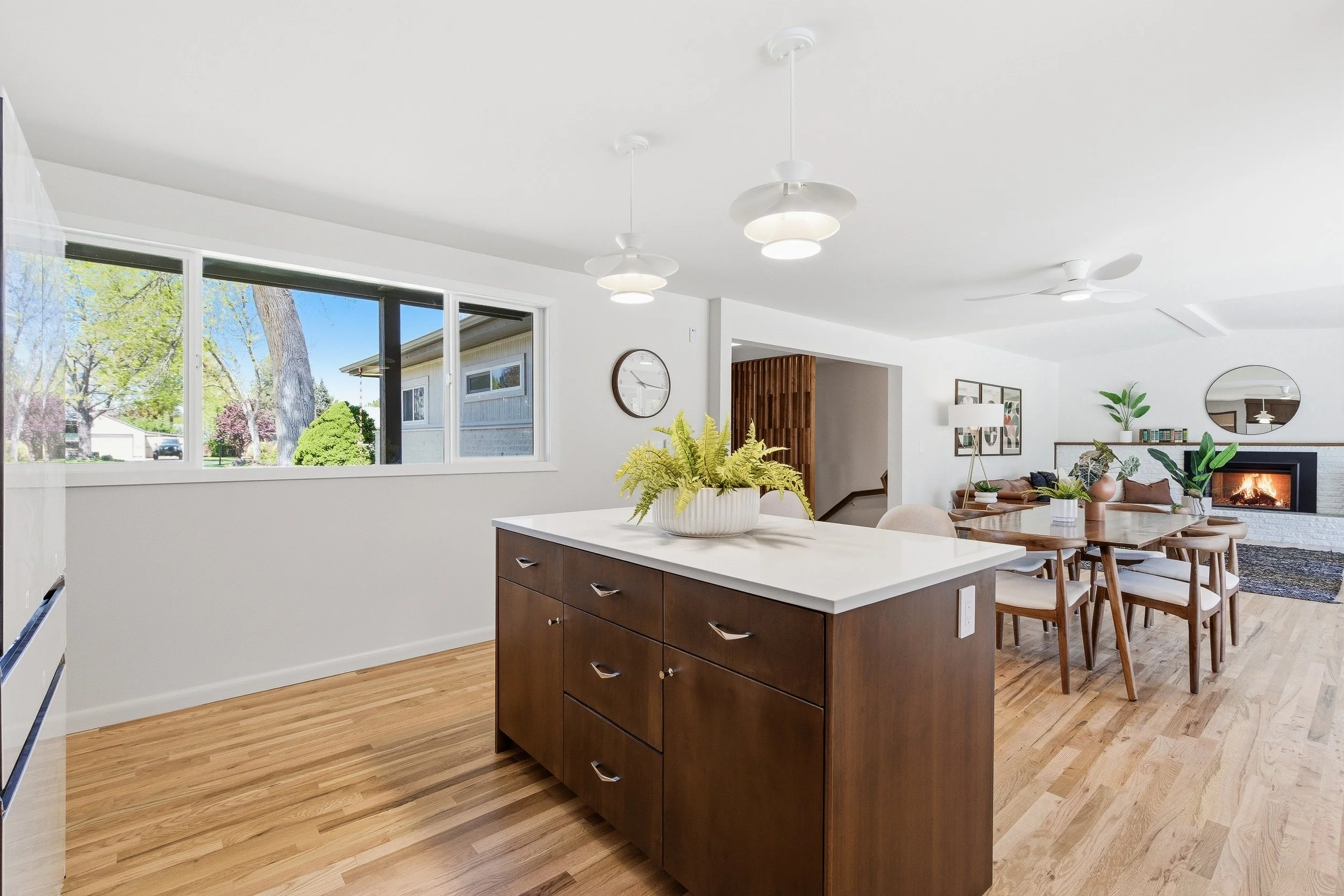 A modern open-concept kitchen and living room area with large windows, wooden flooring, a white island with a potted plant, dining table with chairs, and a fireplace with a mirror above it.