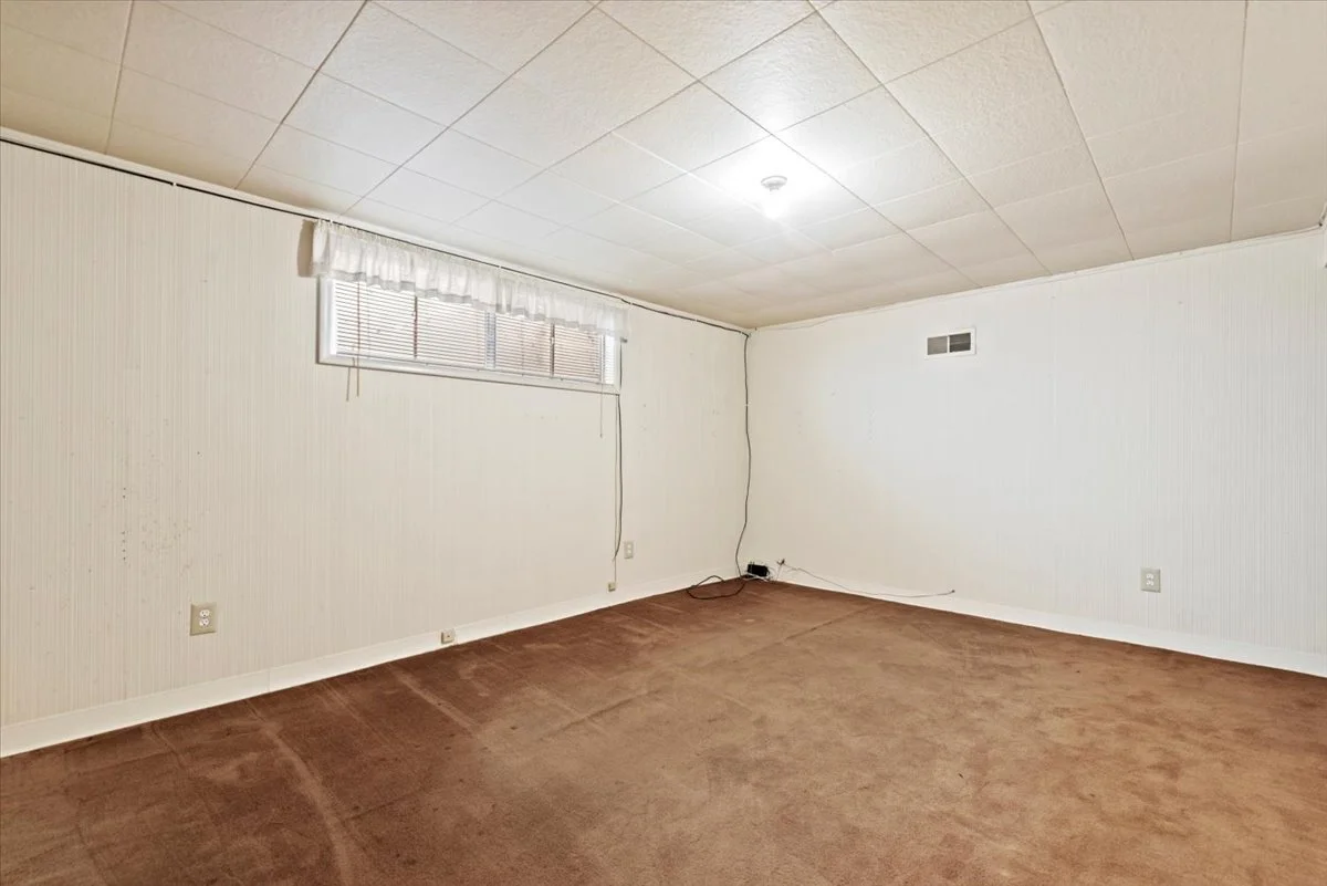 An empty room with beige carpet flooring, white paneled walls, a small window with white curtains, and a ceiling light fixture.