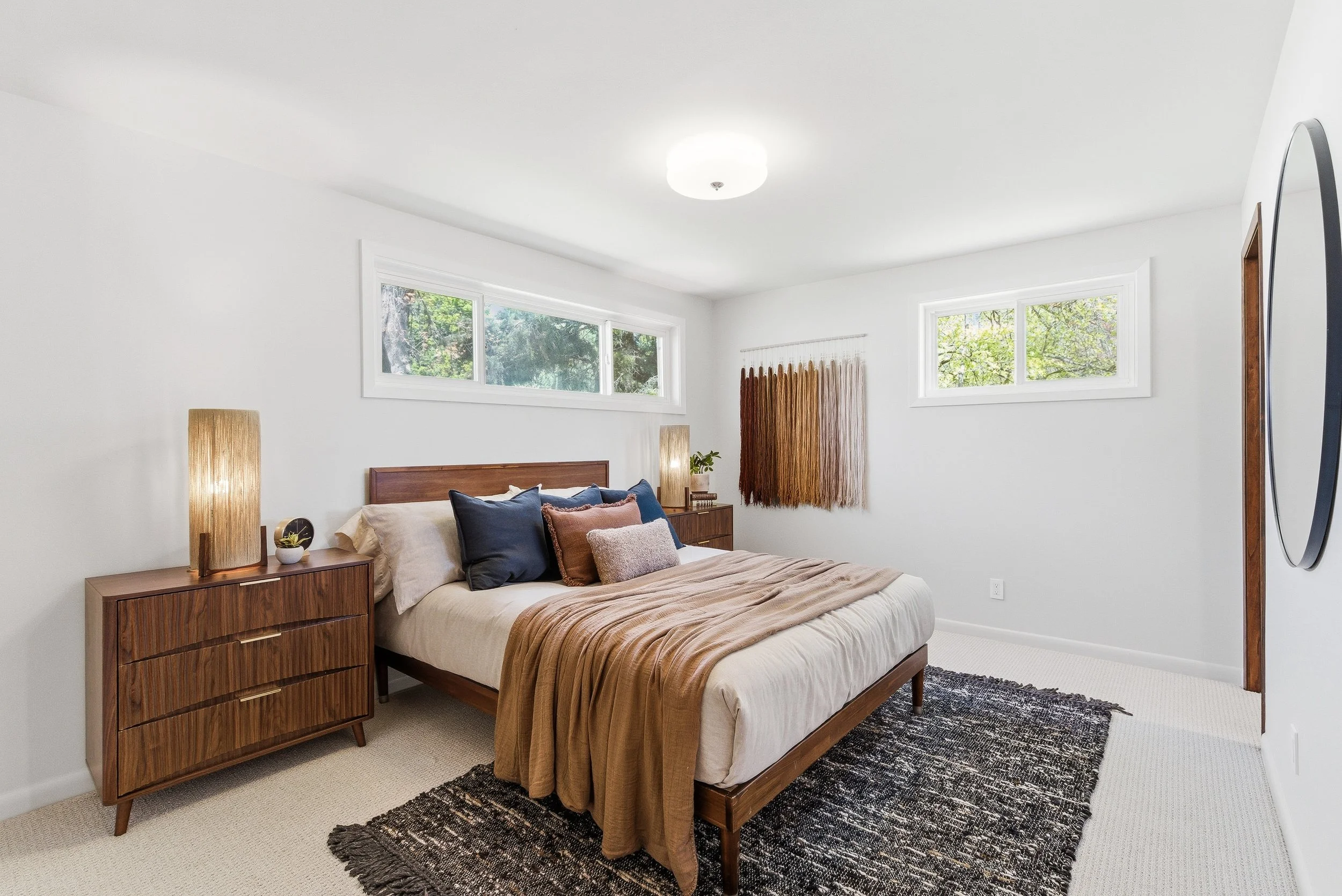 A bedroom with a bed, pillow, and blanket, wooden dresser, two tall lamps, wall art, windows showing trees outside, and a rug.