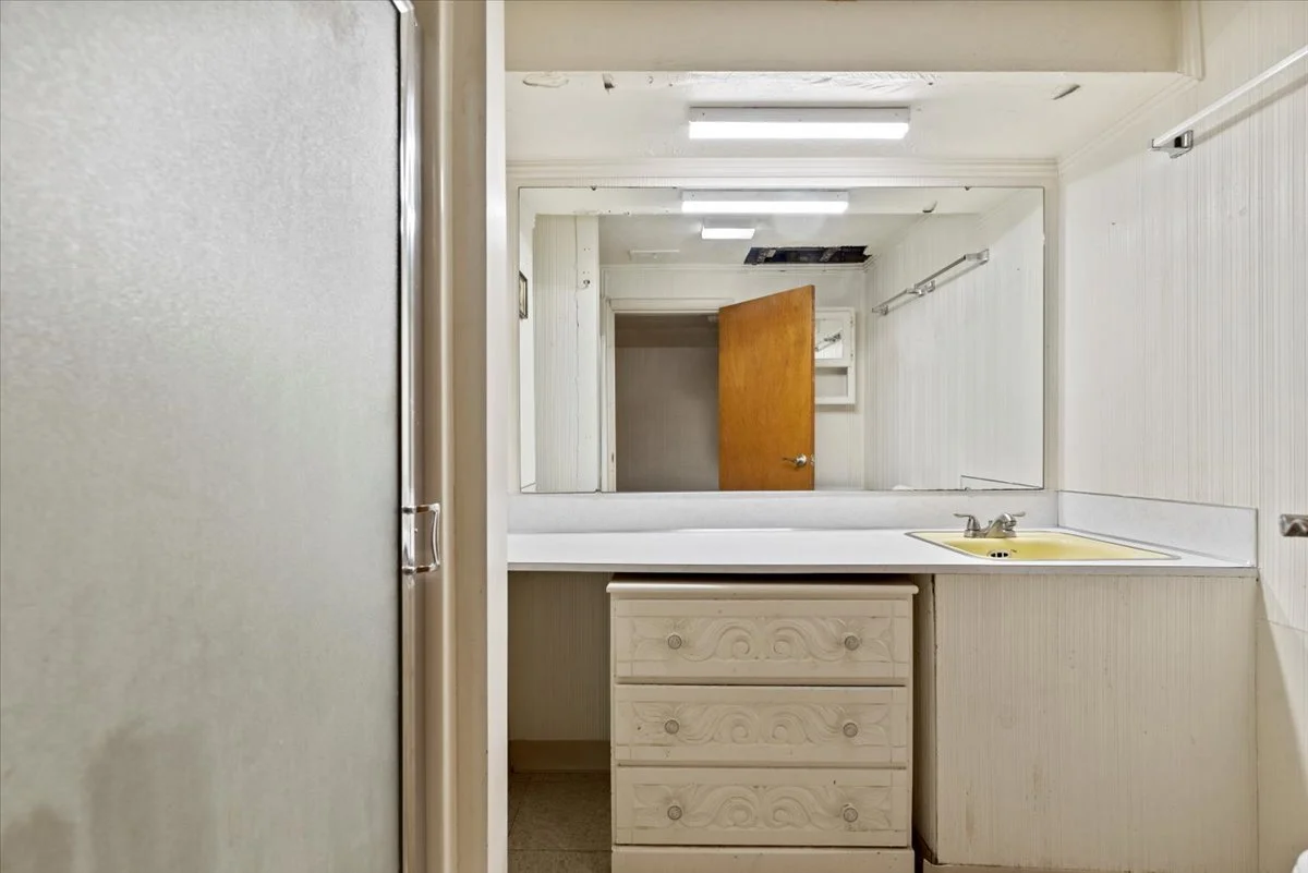 A worn-out bathroom with a large mirror above a yellow sink, a small white dresser with three drawers, and a partially open wooden door leading to another room, with visible ceiling damage and old fluorescent light fixtures.