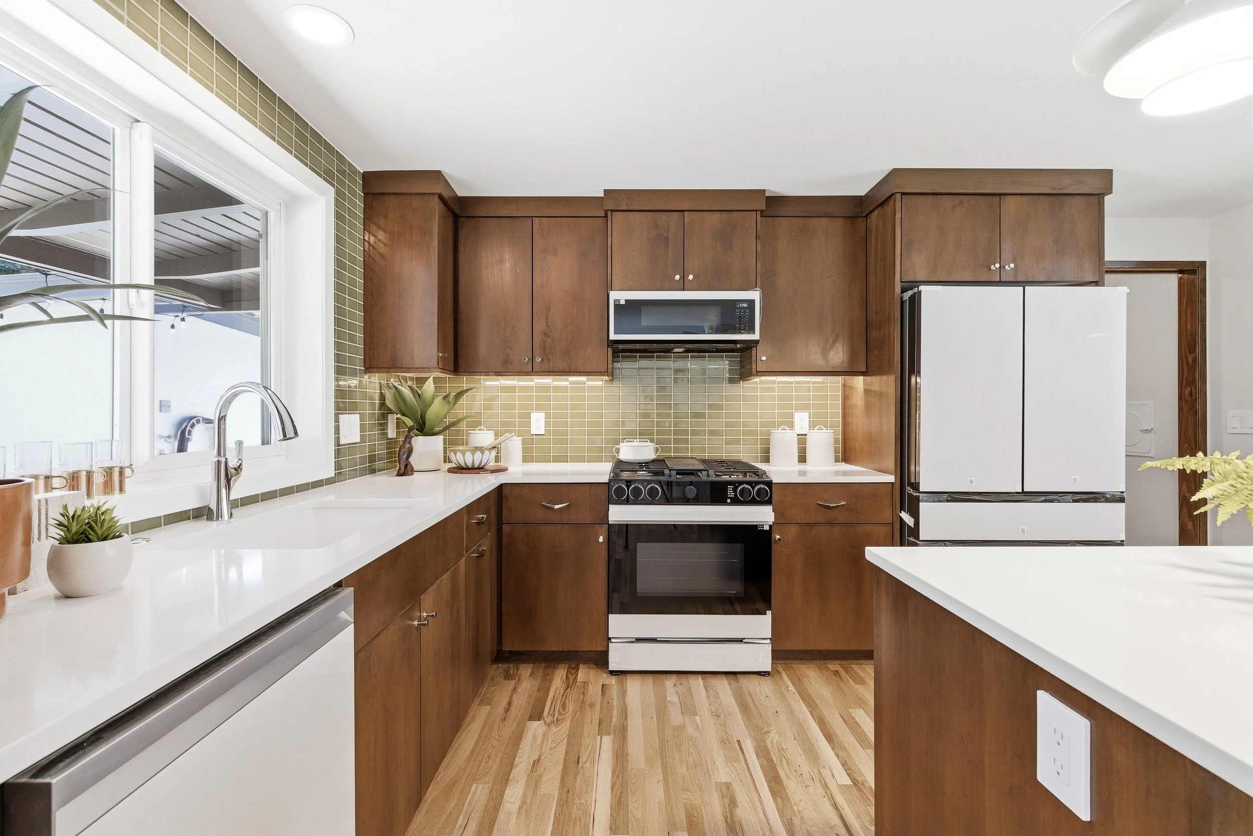 A modern kitchen with wooden cabinets, a white countertop with plants and kitchenware, a window, and appliances including an oven, microwave, and refrigerator.