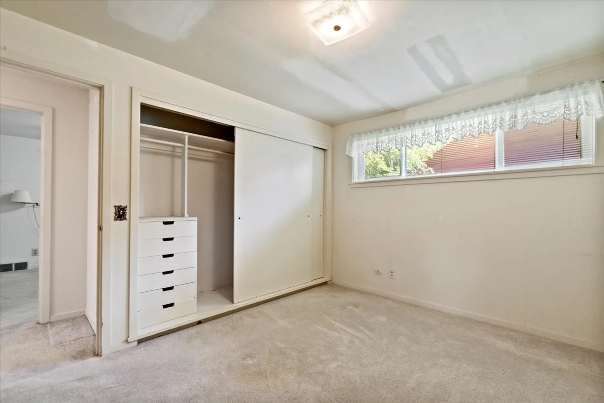 Empty bedroom with a closet, white walls, beige carpet, and a large horizontal window with lace curtains.