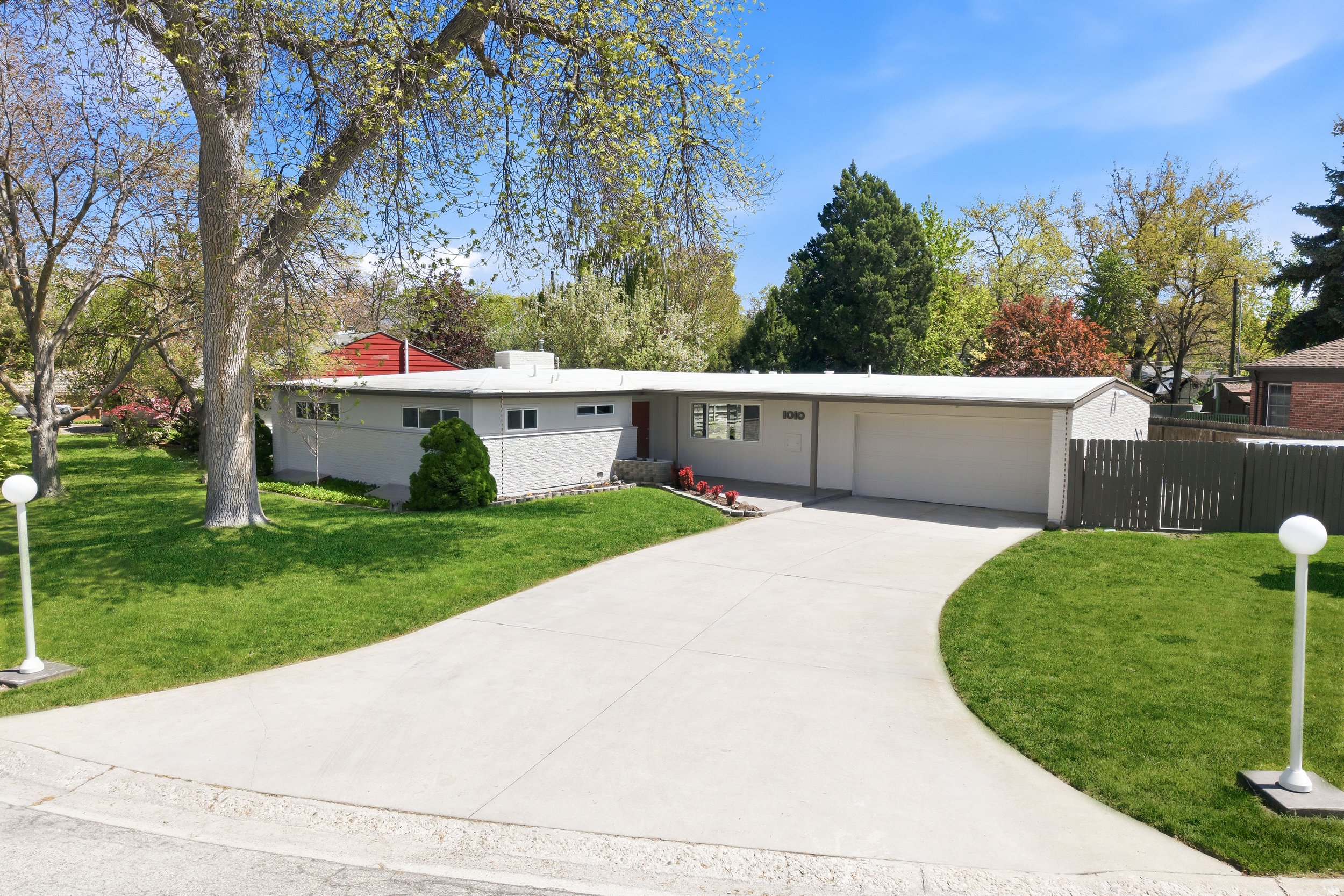 A suburban front yard with green grass, a concrete sidewalk, a white mid-century house with a flat roof, and a detached garage. Several large trees with budding leaves are in the yard and background, with a bright blue sky overhead.
