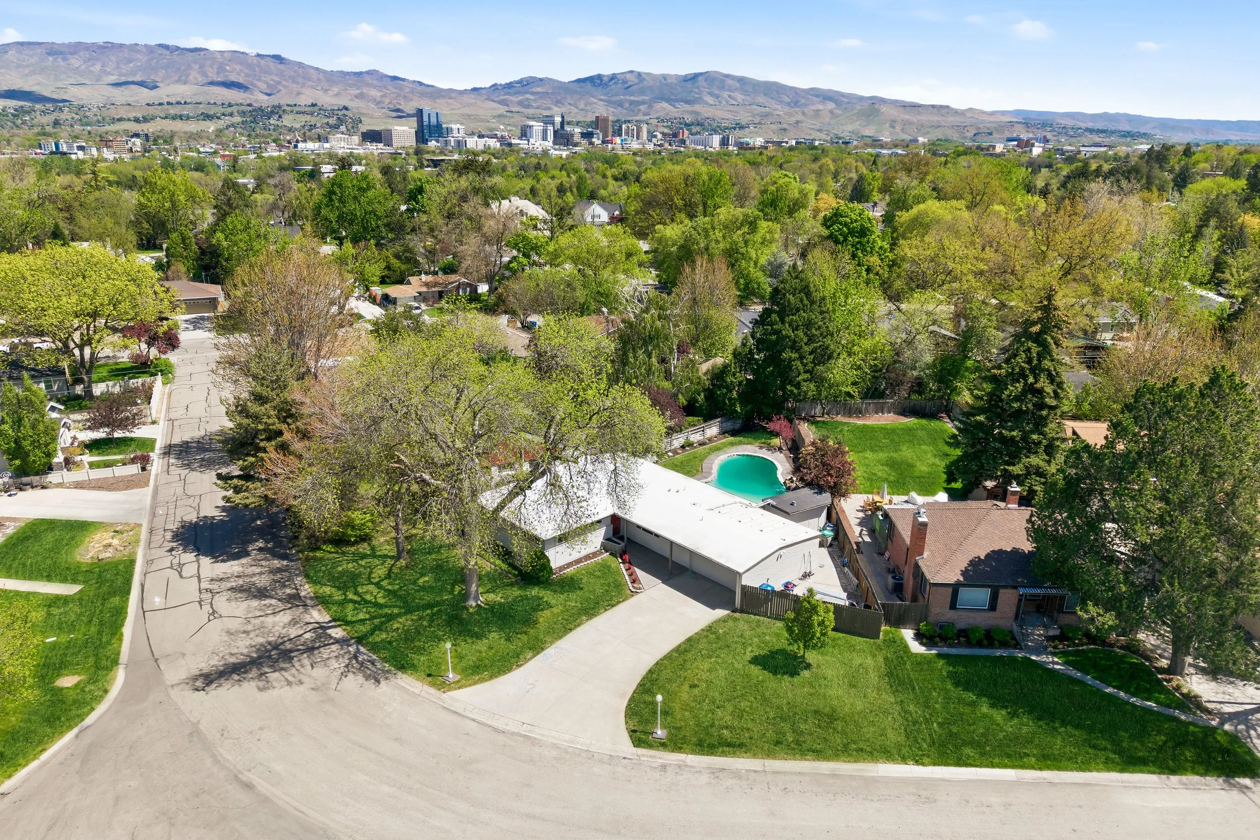 Aerial view of a residential backyard with a swimming pool, surrounded by trees and houses, with the city skyline and green mountains in the distance under a clear blue sky.