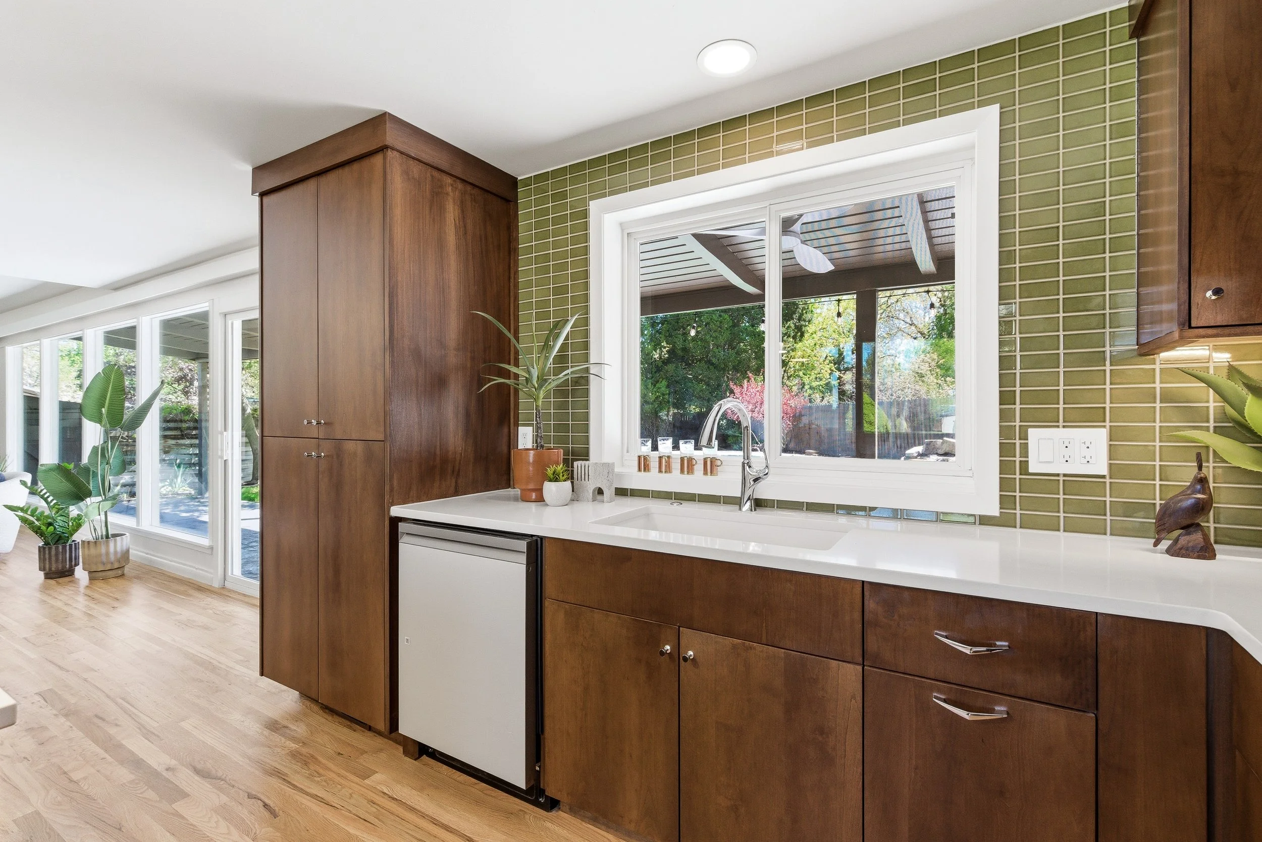 Modern kitchen with wood cabinets, a white countertop, green tile backsplash, and large window overlooking a backyard with trees and patio. Potted plants on the counter and a small wooden bird sculpture.