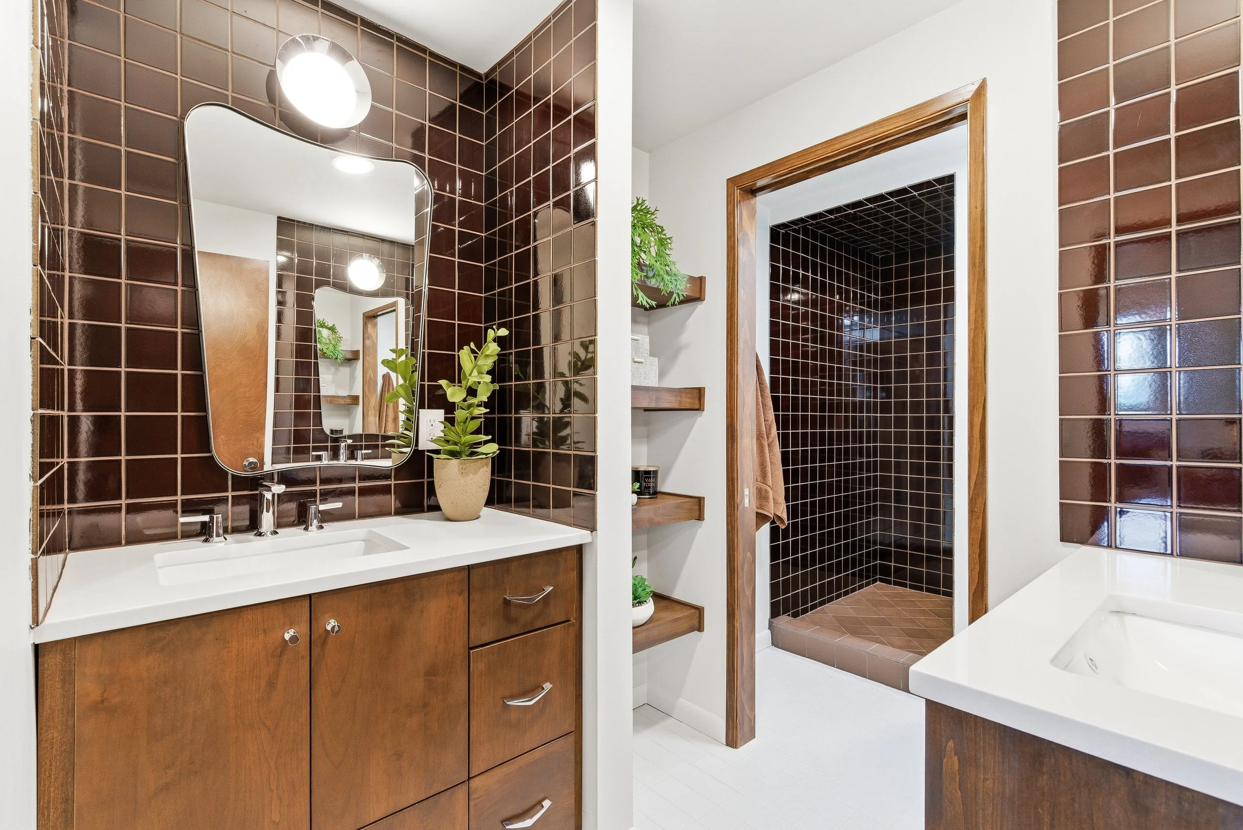 Bathroom with brown tiled walls, a mirror, wooden vanity with drawers, and a sink. A small plant on the countertop, shelving with decorations, and a separate shower area with dark tiles and a wooden frame.