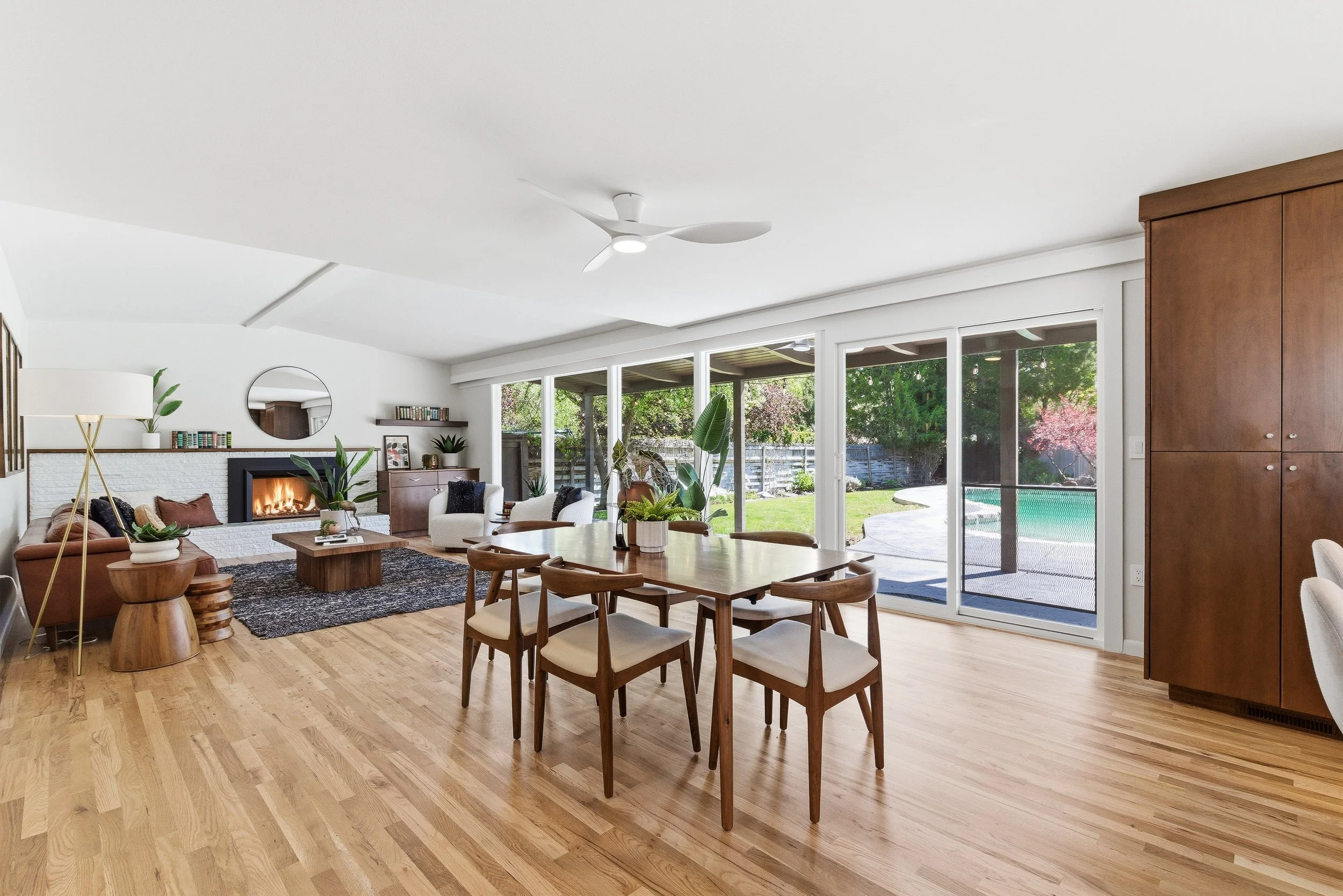A modern living room with large glass sliding doors showing greenery outside. The space features wooden flooring, a white ceiling fan, a fireplace, and various seating options including a sofa and armchairs. There's a wooden dining table with chairs,