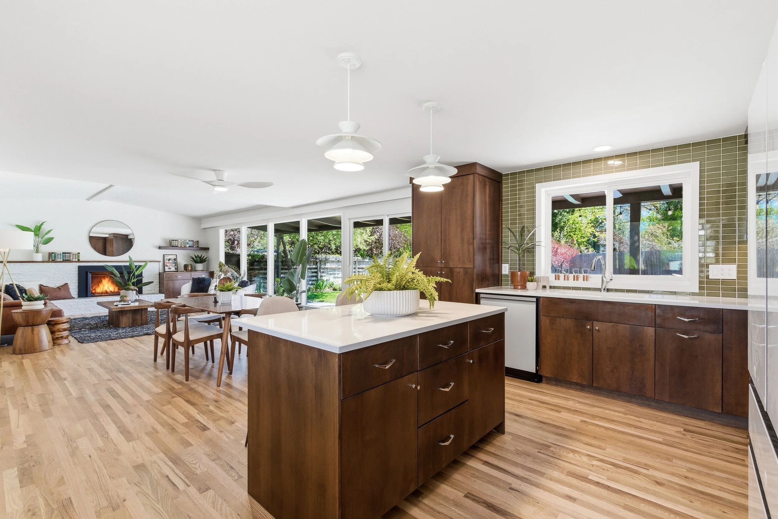 Open-concept kitchen and living room with wooden floors, white walls, large windows showing green trees outside, and modern white pendant lights.