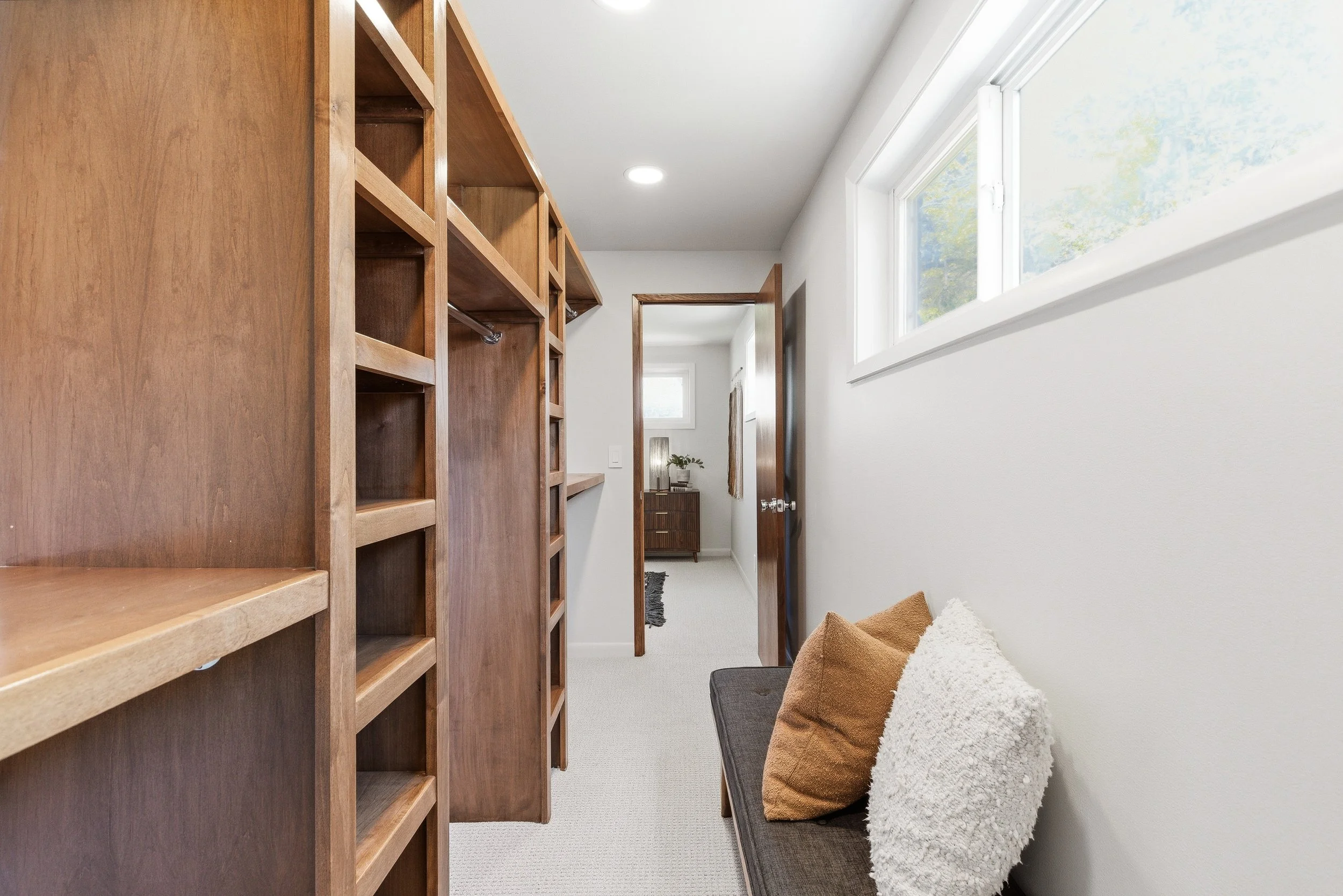 A narrow hallway with a wooden bookshelf on the left, windows on the right, and a small seating area with pillows at the bottom right. A door at the end of the hall is open, leading into another room.