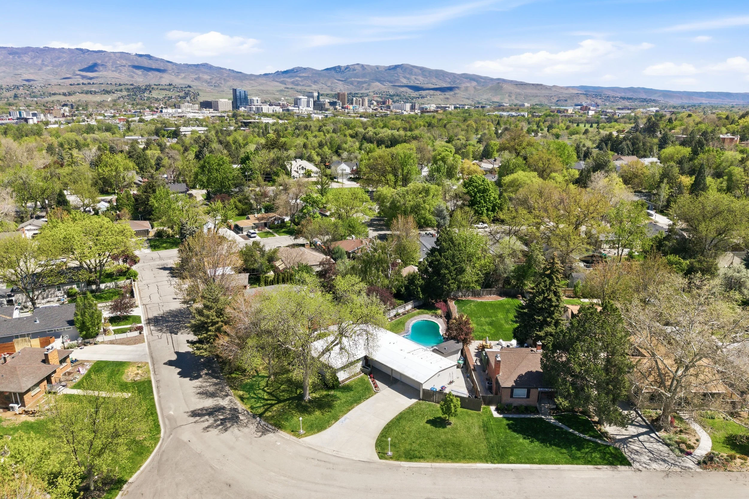 Aerial view of a suburban neighborhood with houses, trees, and a backyard pool, with a cityscape and mountains in the background under a partly cloudy sky.