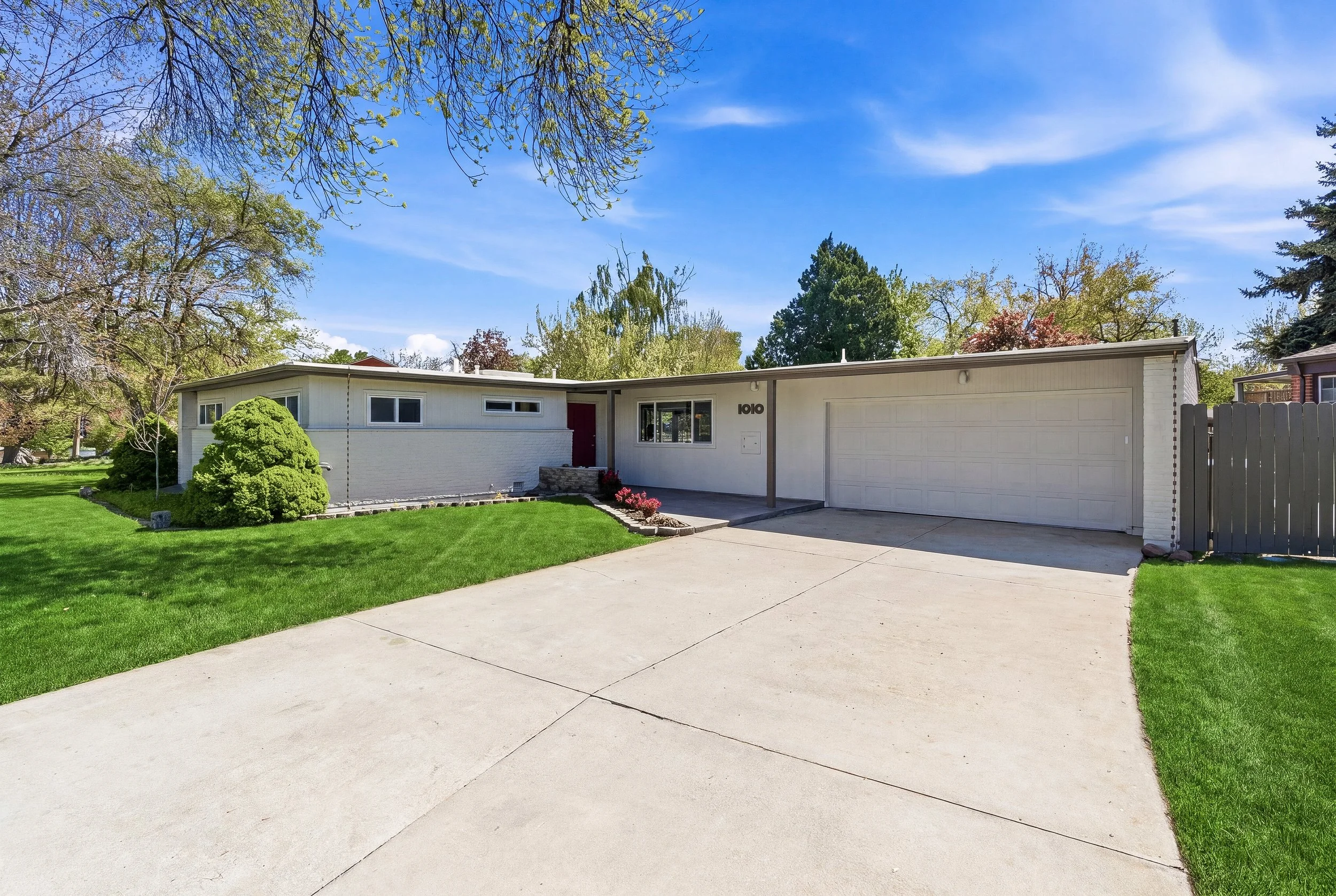Front view of a single-story house with a white exterior, red door, and gray roof, surrounded by a green lawn, trees, and a clear blue sky.