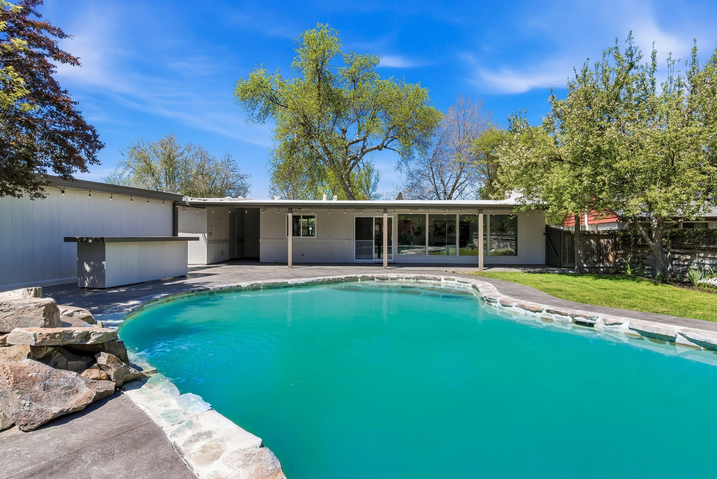 Backyard with a swimming pool, surrounded by a patio, trees, and a modern house with large glass windows, under a clear blue sky.