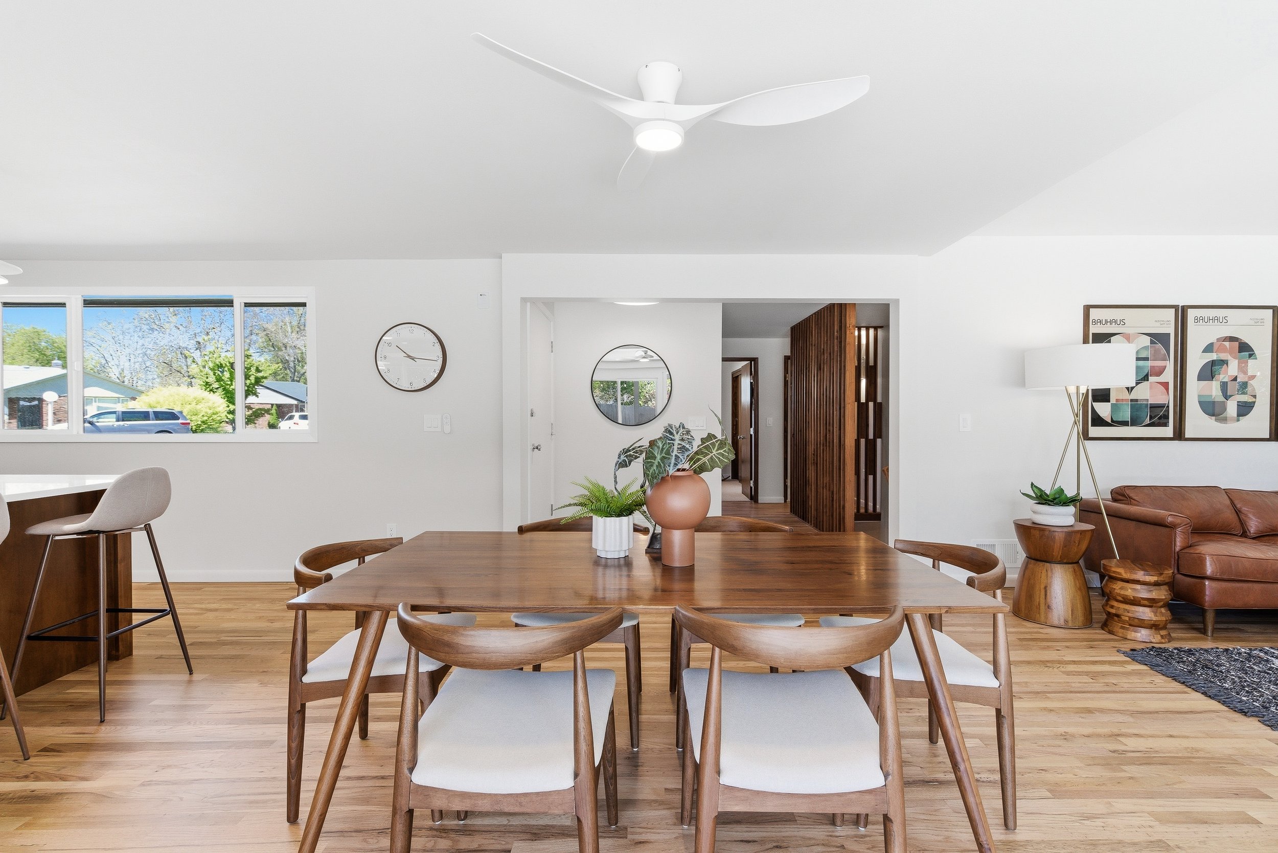 A bright, modern dining room with a wooden table and six matching chairs, a white ceiling fan, linear windows showing a sunny outdoor scene, and art prints on the wall, with some inside plants in the room.