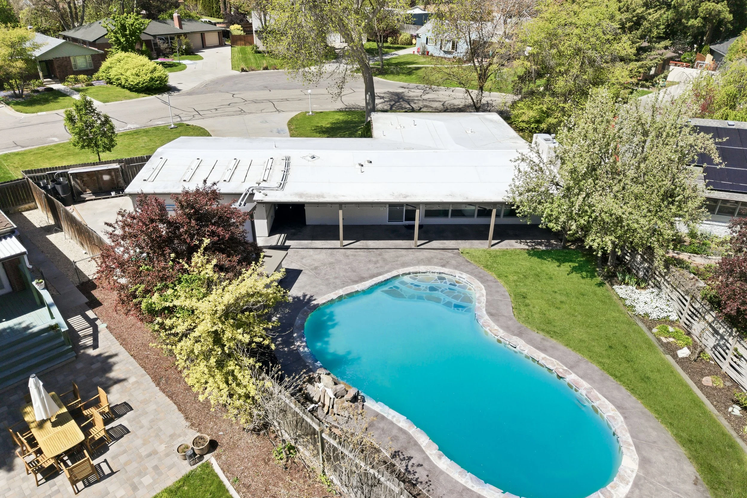 Aerial view of a residential backyard with a swimming pool, surrounded by green grass, trees, and neighboring houses. A patio area with outdoor furniture is visible on the left. The house has a flat roof and there are solar panels on a neighboring ro