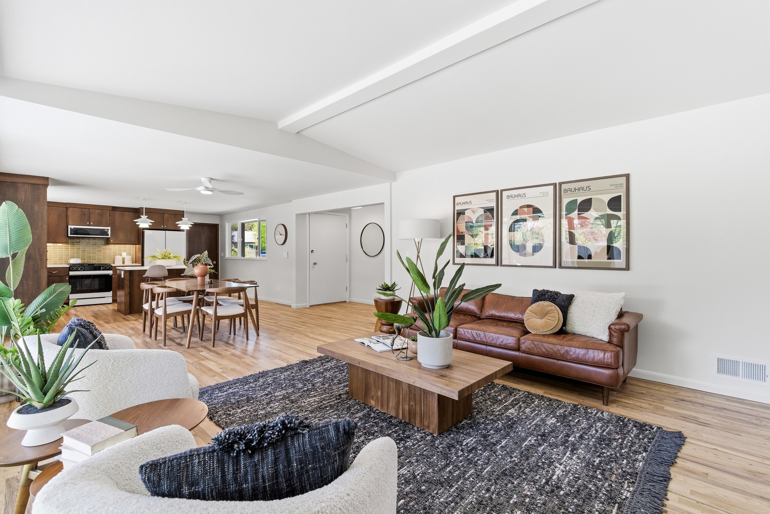 Interior of a modern living room with white walls, wooden floors, a brown leather sofa, a white armchair, a wooden coffee table with potted plants, framed abstract artwork on the wall, and part of the kitchen with wooden cabinets in the background.