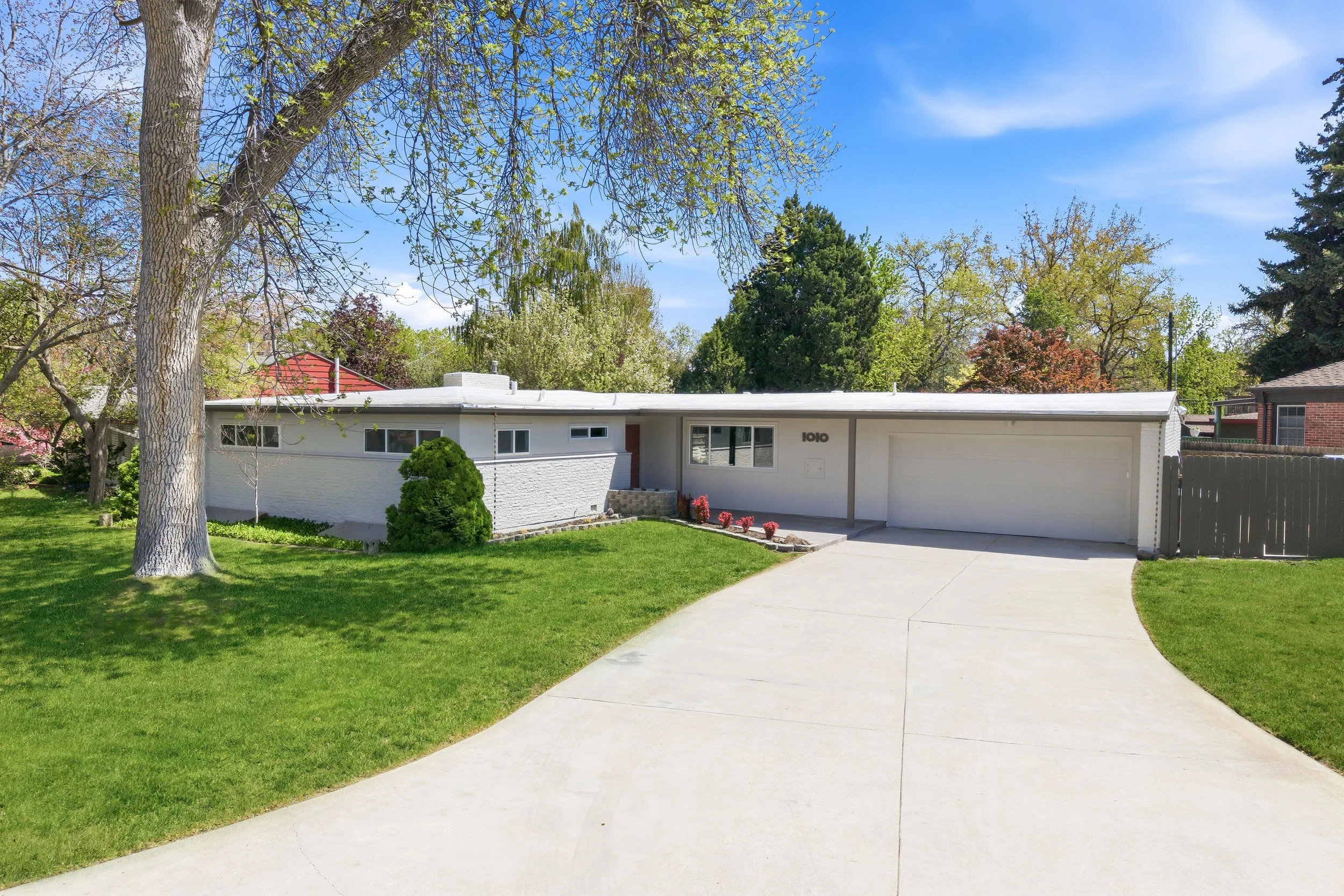 A white mid-century modern house with a flat roof and a one-car garage, surrounded by green grass and trees, on a sunny day with a clear blue sky.