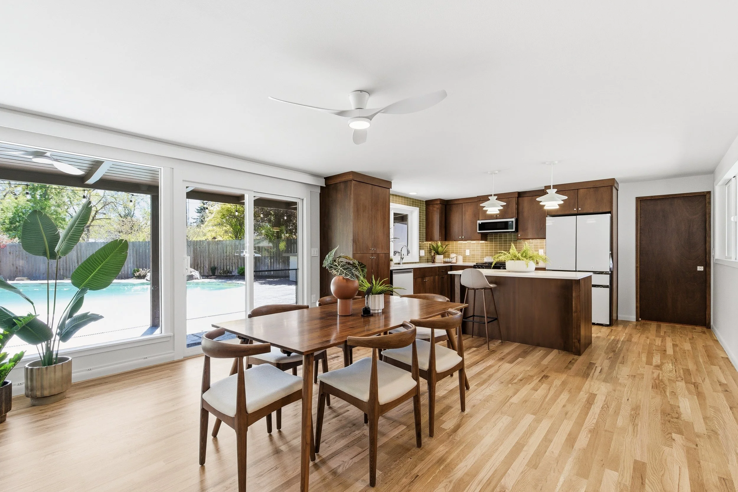 Bright open kitchen and dining area with wooden flooring, large windows, a wooden dining table with chairs, and modern kitchen cabinets with pendant lights.