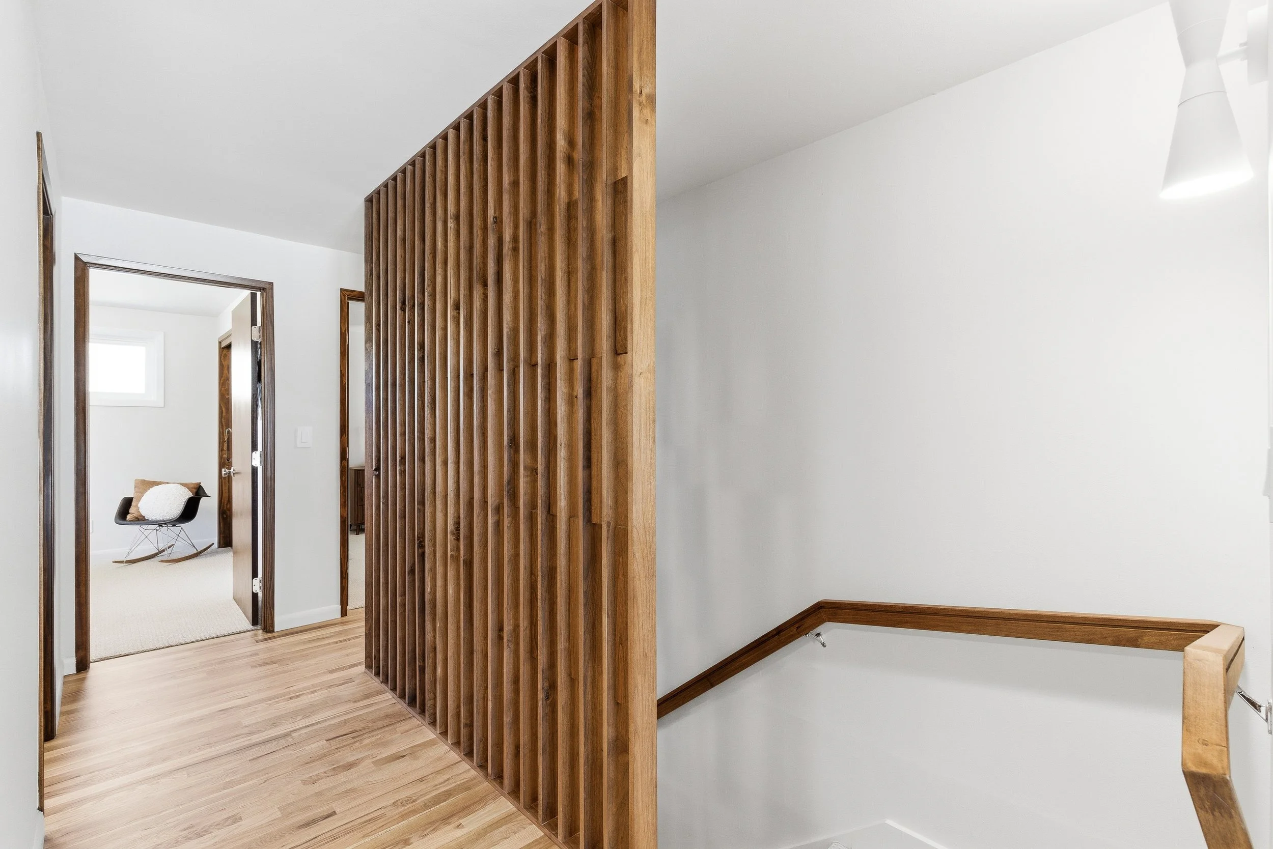 Interior view of a modern home with a wooden slatted room divider, white walls, wooden trim, and a staircase with a banister. A doorway leads to a room with a chair and window.