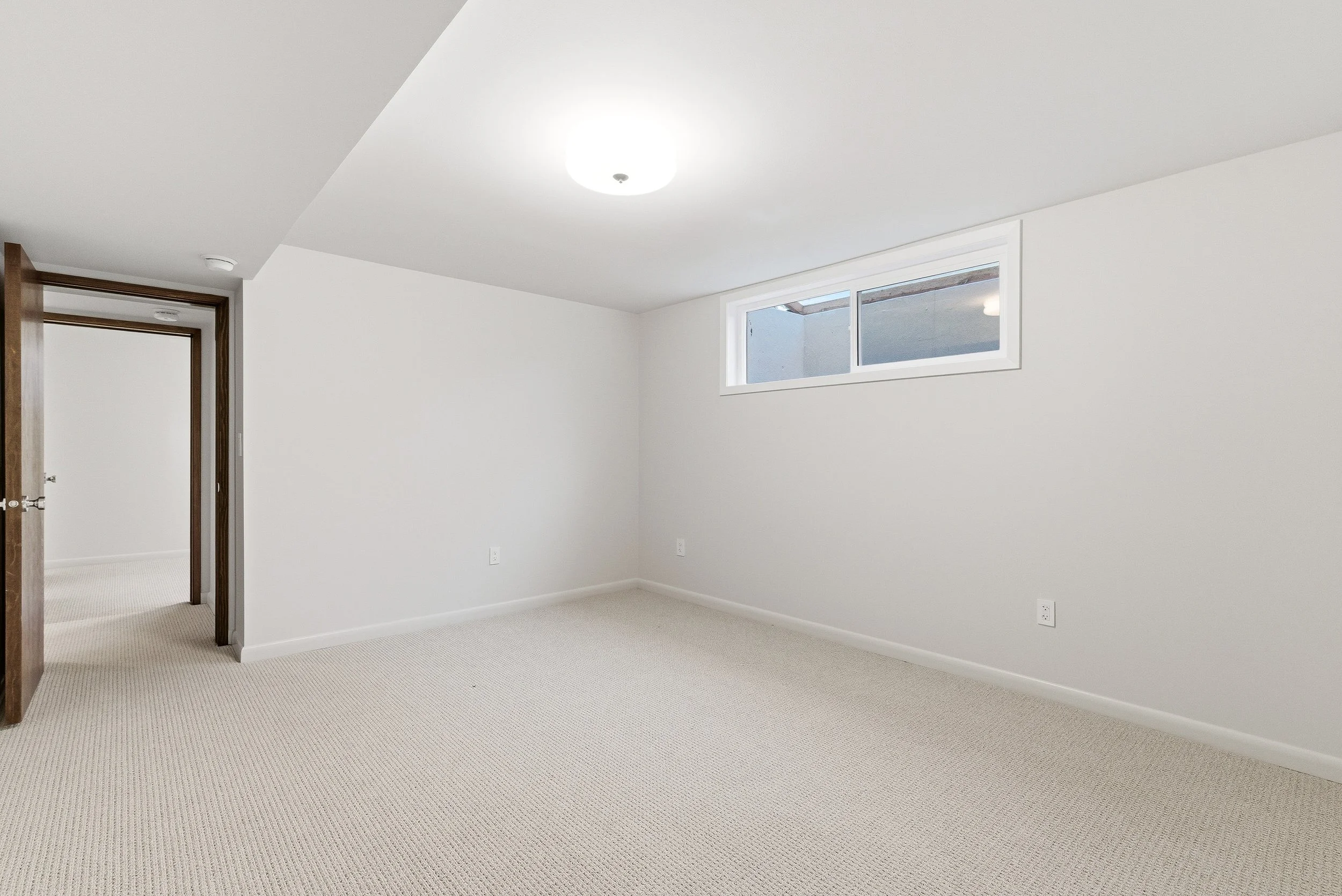 Empty room with white walls, beige carpet, a small window, and a ceiling light fixture.