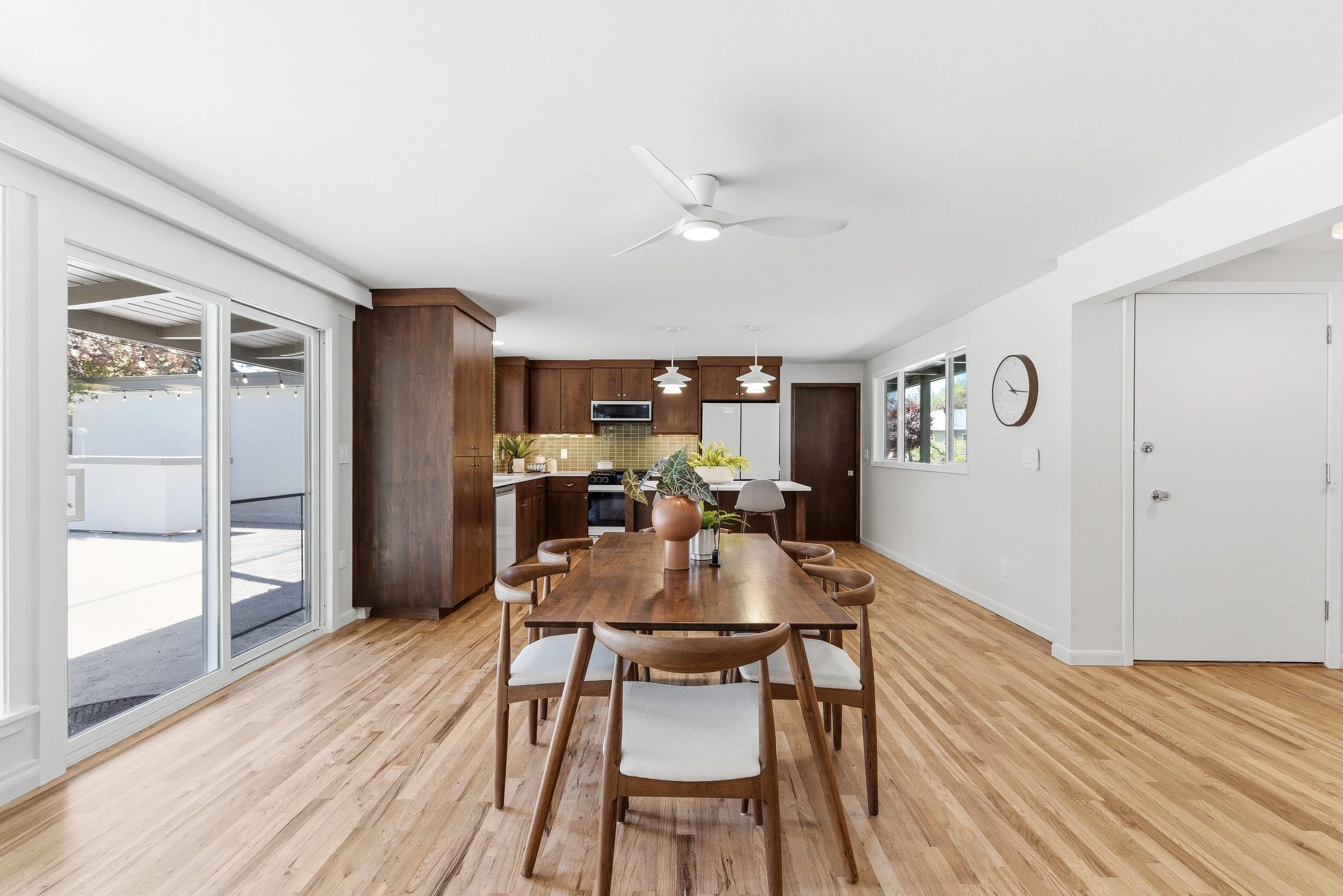 Modern open-concept kitchen and dining area with wooden flooring, white walls, large sliding glass doors leading to outdoor space, ceiling fan, and minimalist decor.