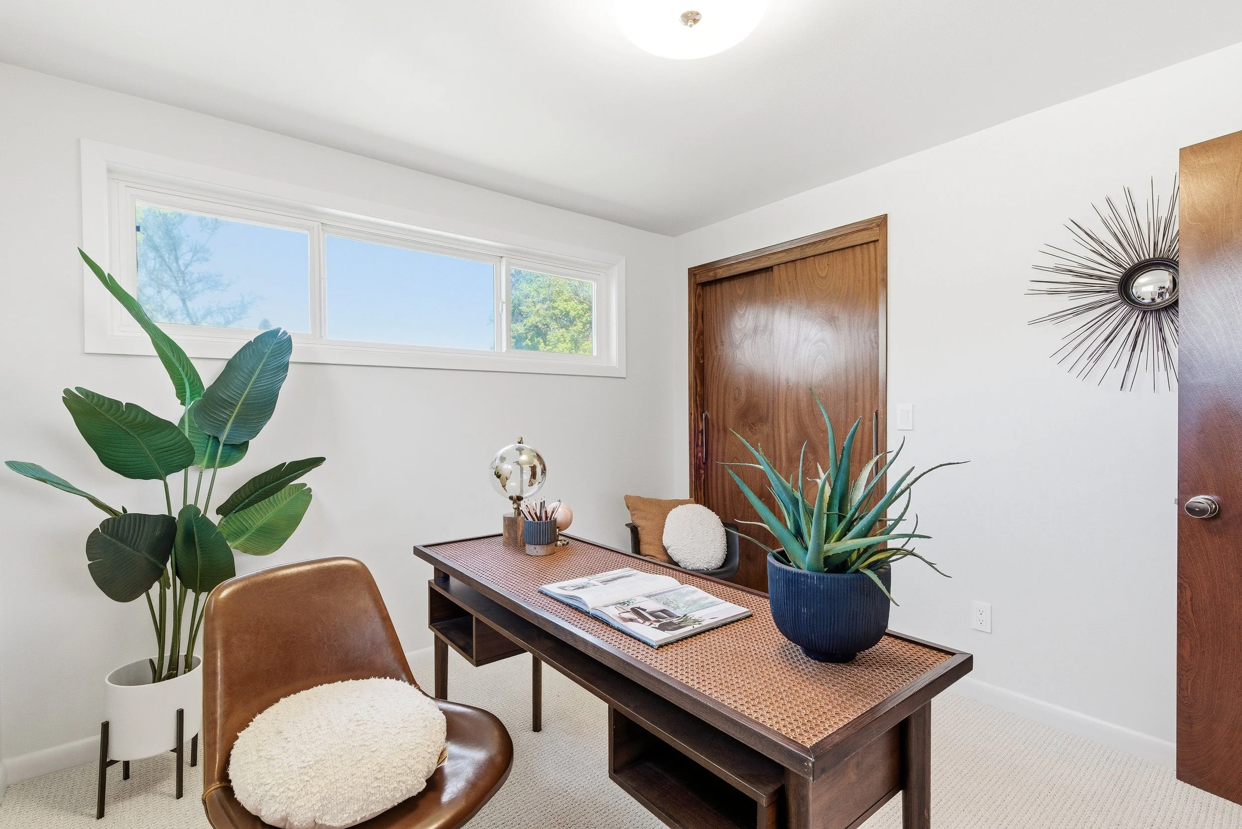 Interior of a modern home office with white walls, a large window, a wooden door, a desk with magazines, plants, and decorative items, and brown chairs.