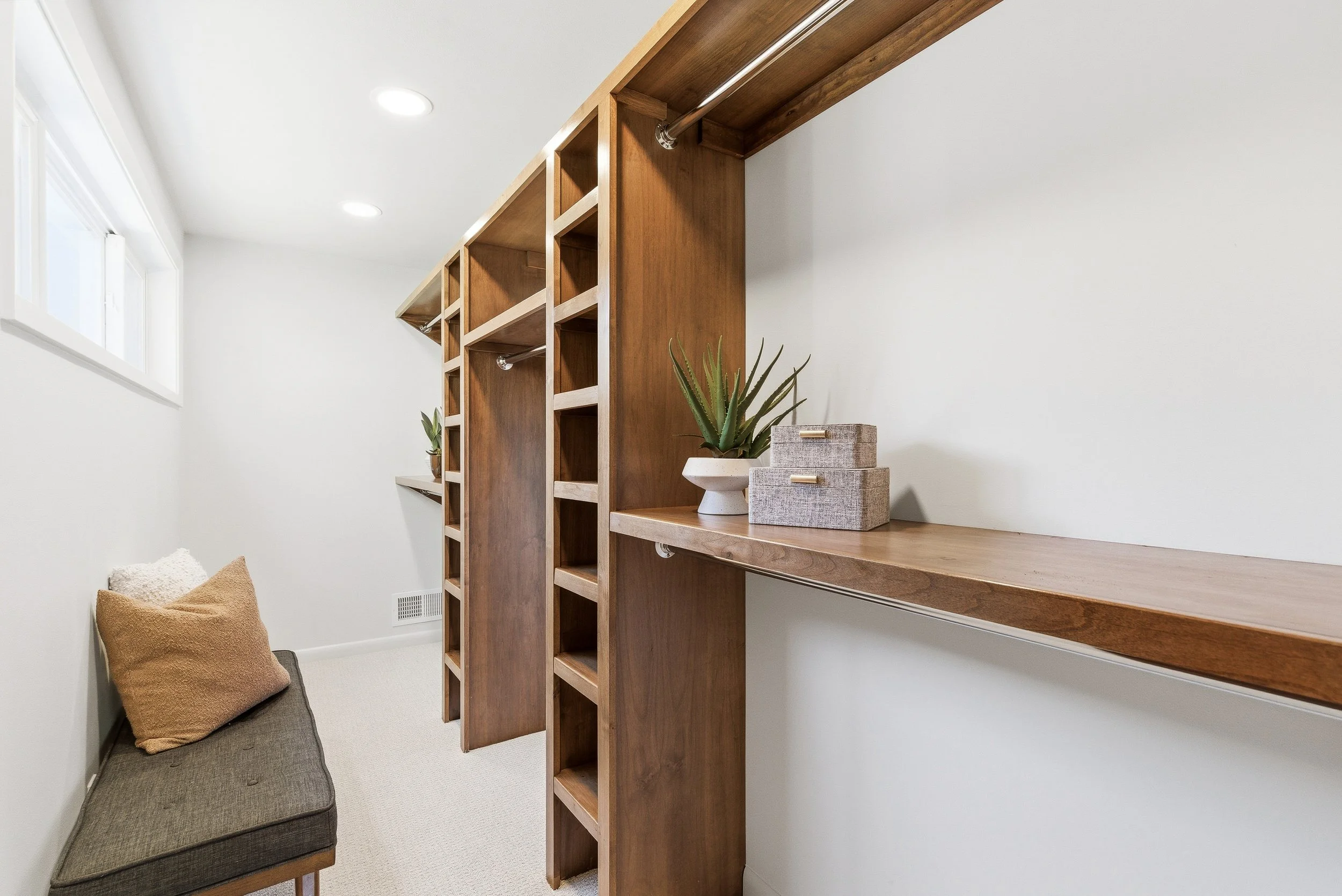 A corner of a room with a built-in wooden shelving unit, a small gray couch with a cushion, a plant in a white pot, and a window letting in natural light.
