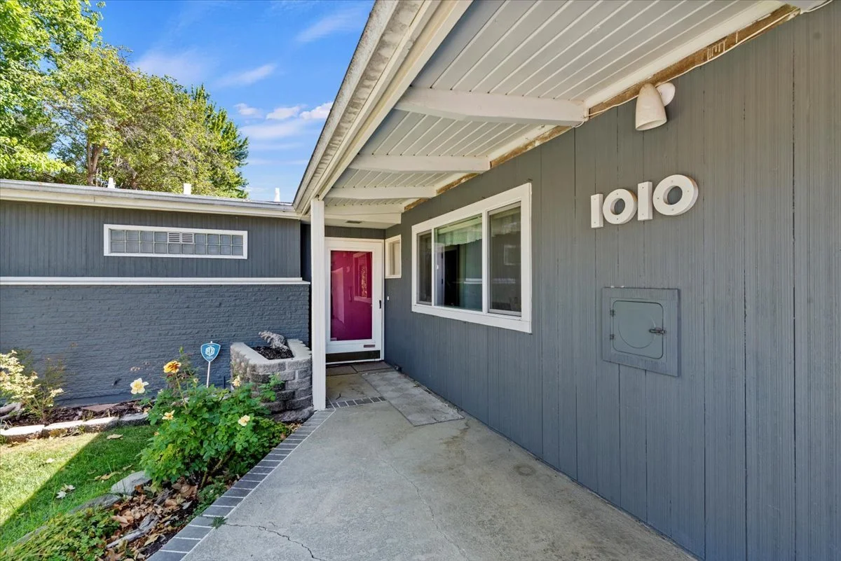 Front entrance of a house with a gray exterior, a purple door, a window, a small garden with flowers, and house numbers 1010 mounted on the wall.