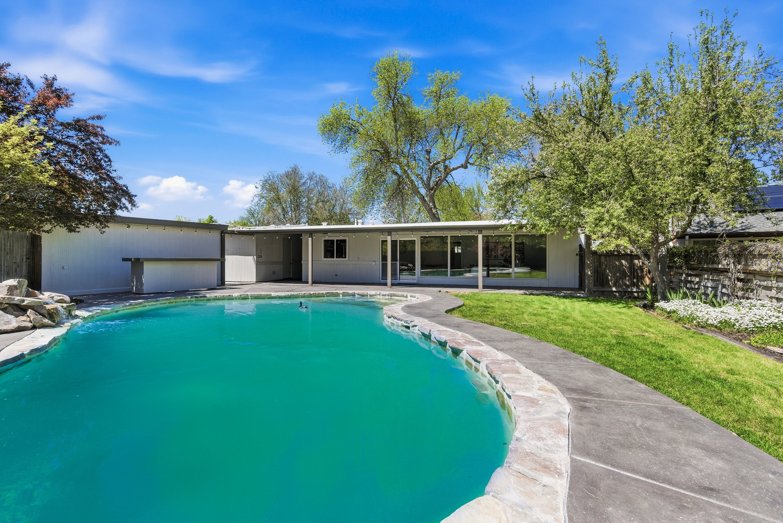 A backyard swimming pool with clear blue water, surrounded by a stone border and a grassy lawn, with trees and a white house in the background under a bright blue sky with some clouds.