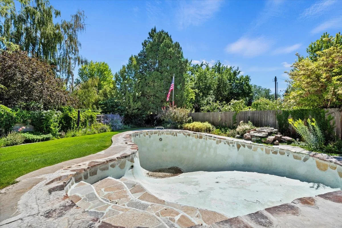 Empty backyard swimming pool with stone edging, surrounded by green lawn and trees under a blue sky.