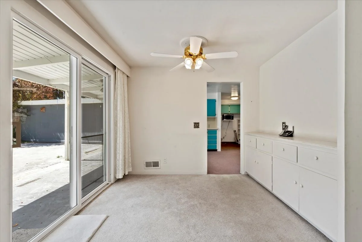 Empty room with sliding glass door leading outside, white carpet, built-in white cabinet, ceiling fan with lights, visible kitchen through doorway