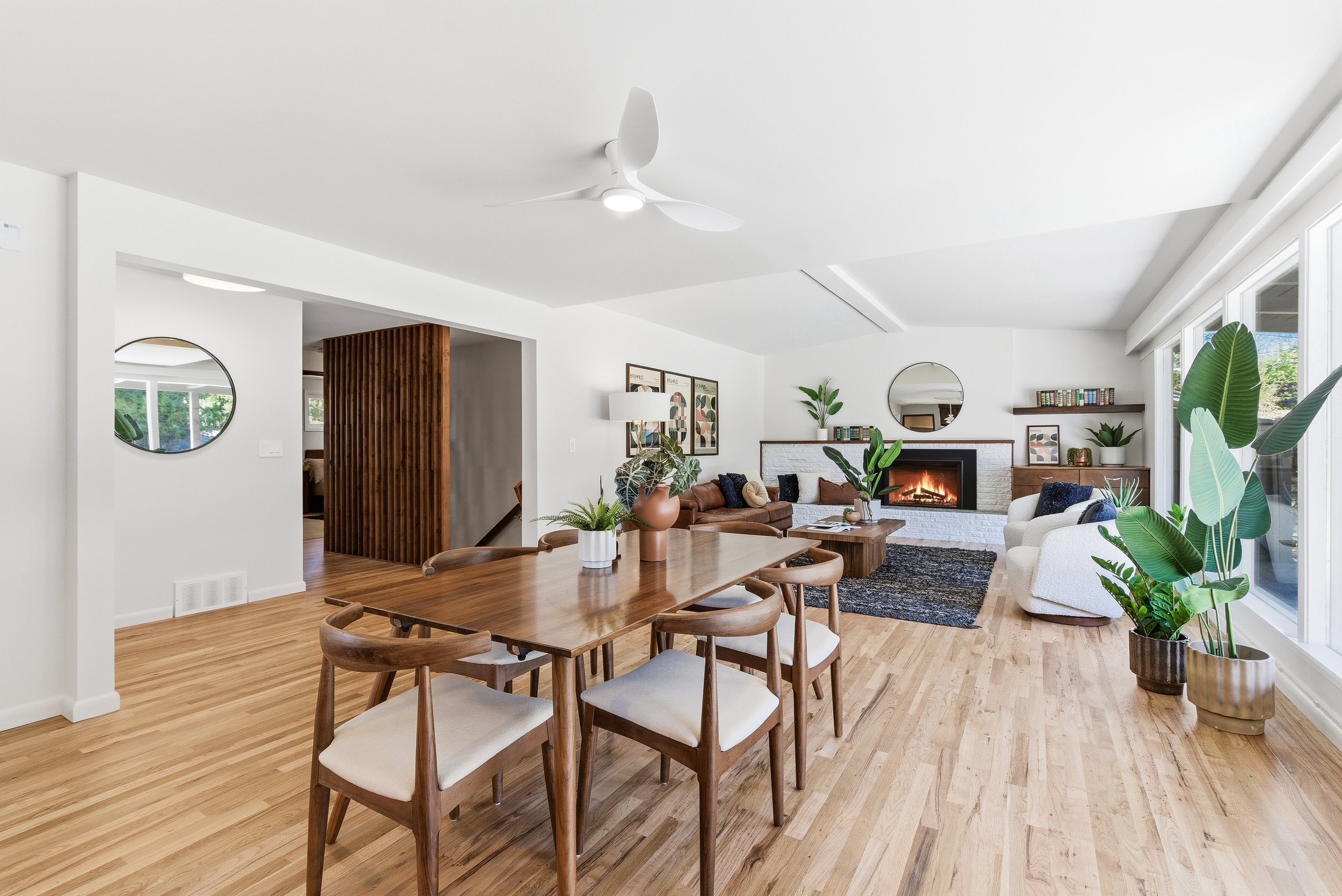 Bright, modern living and dining room with white walls and ceiling, light wooden flooring, a wooden dining table with six matching chairs, a sofa set near a fireplace, large windows with plants, and artwork on the walls.