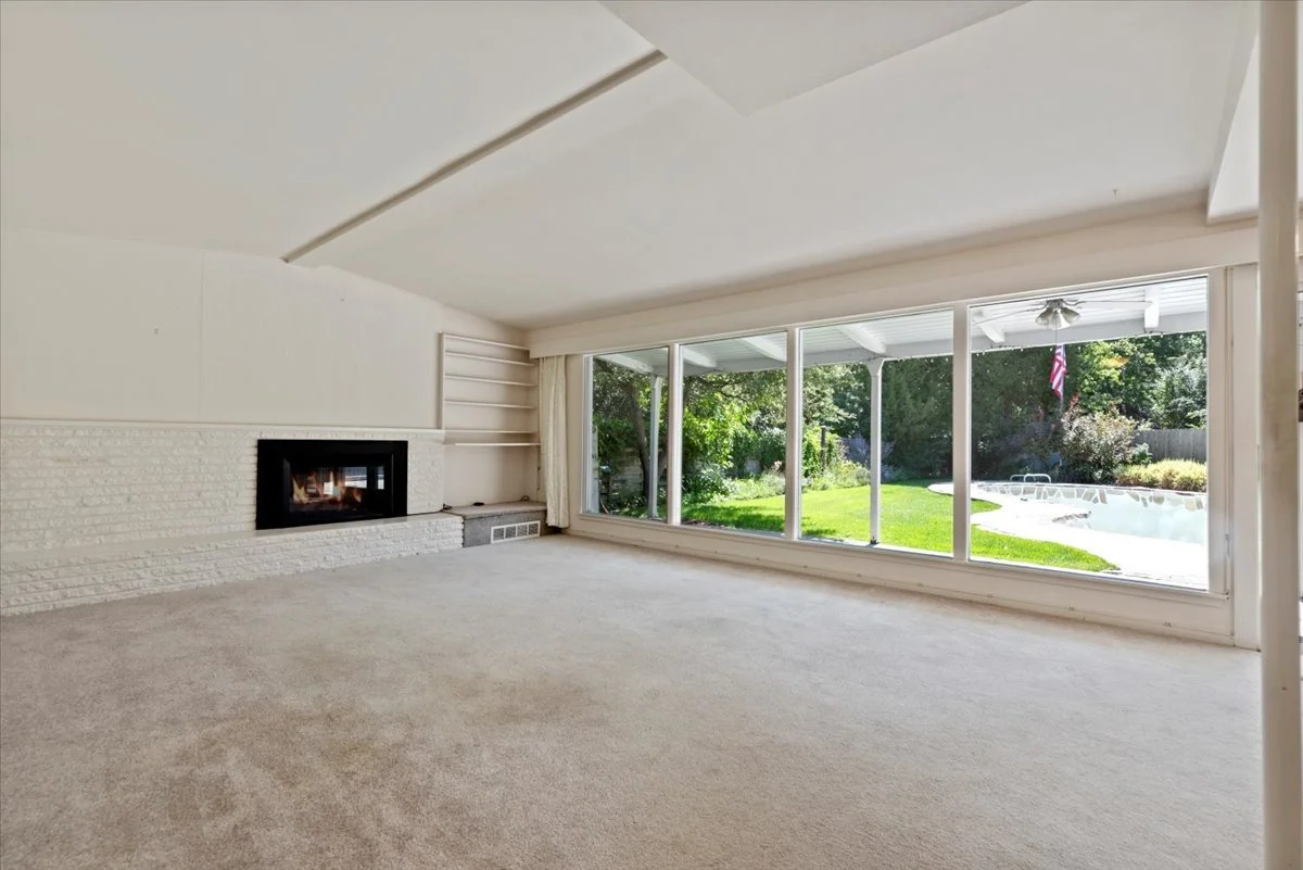 Empty living room with large sliding glass doors overlooking a backyard with a swimming pool and green trees.