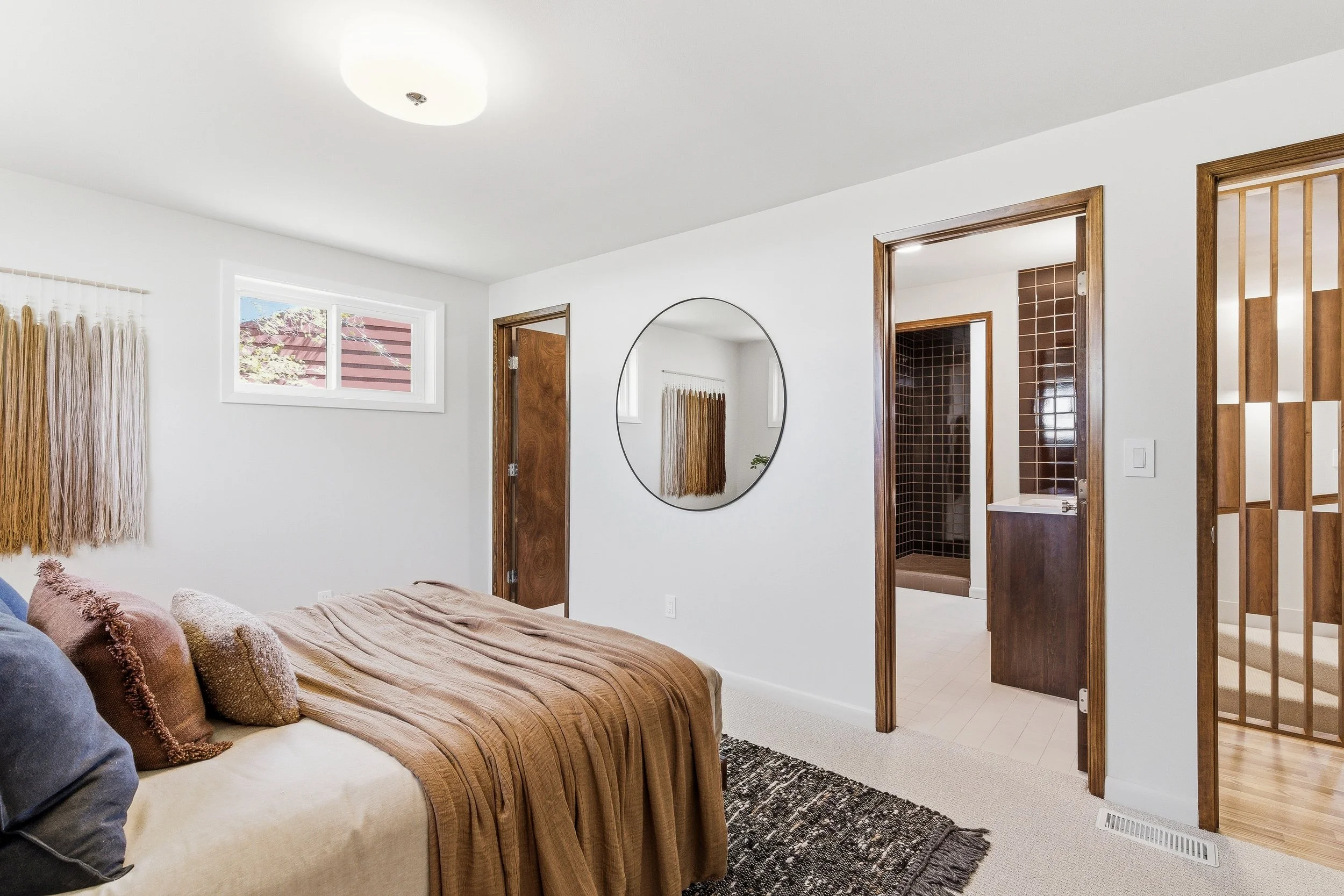 A bedroom with a bed, pillows, and a window, connected to a bathroom with a shower, a mirror, and wooden door frames.