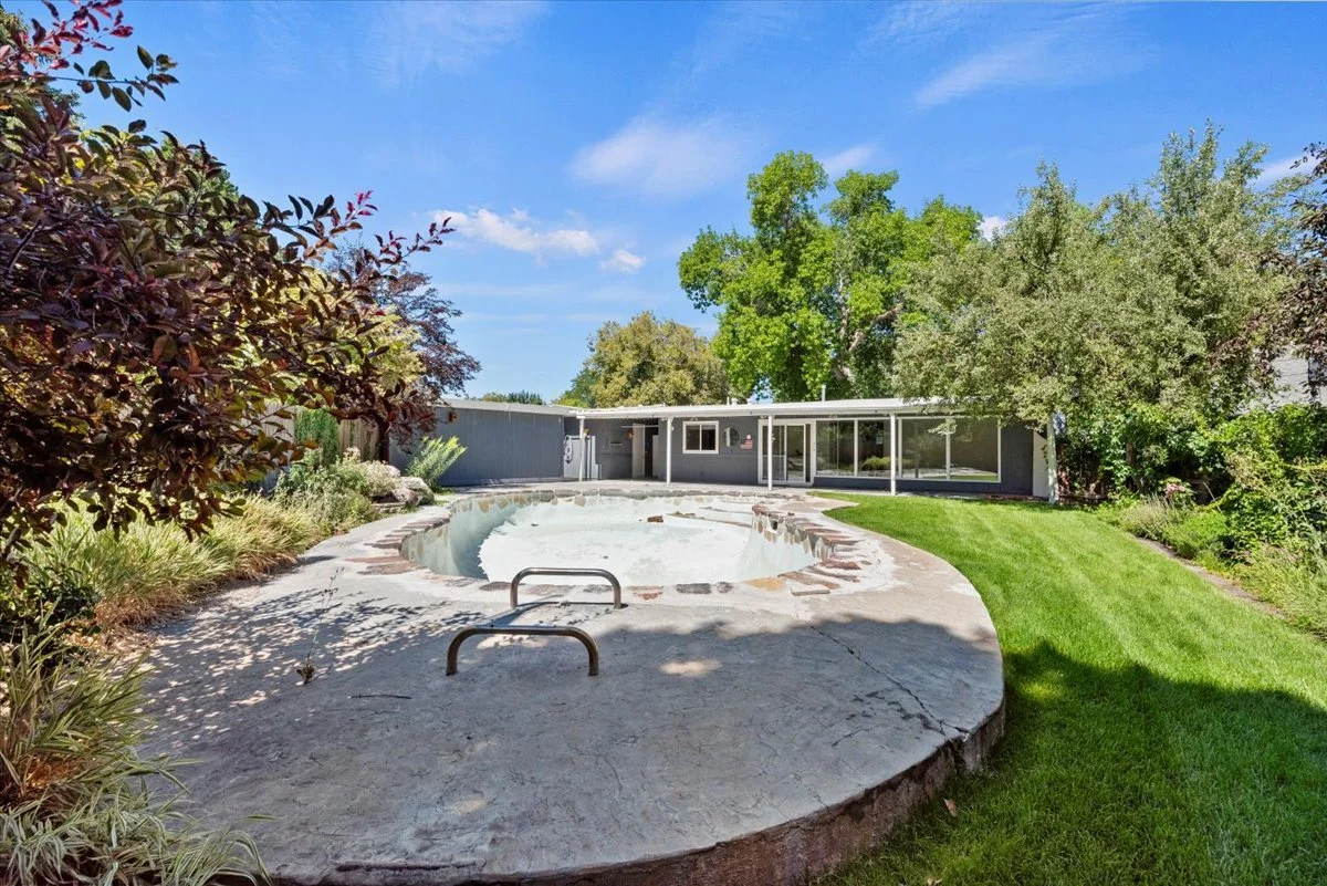 Front yard of a house with a round empty pool, green lawn, trees, and a gray house with large windows and sliding doors.