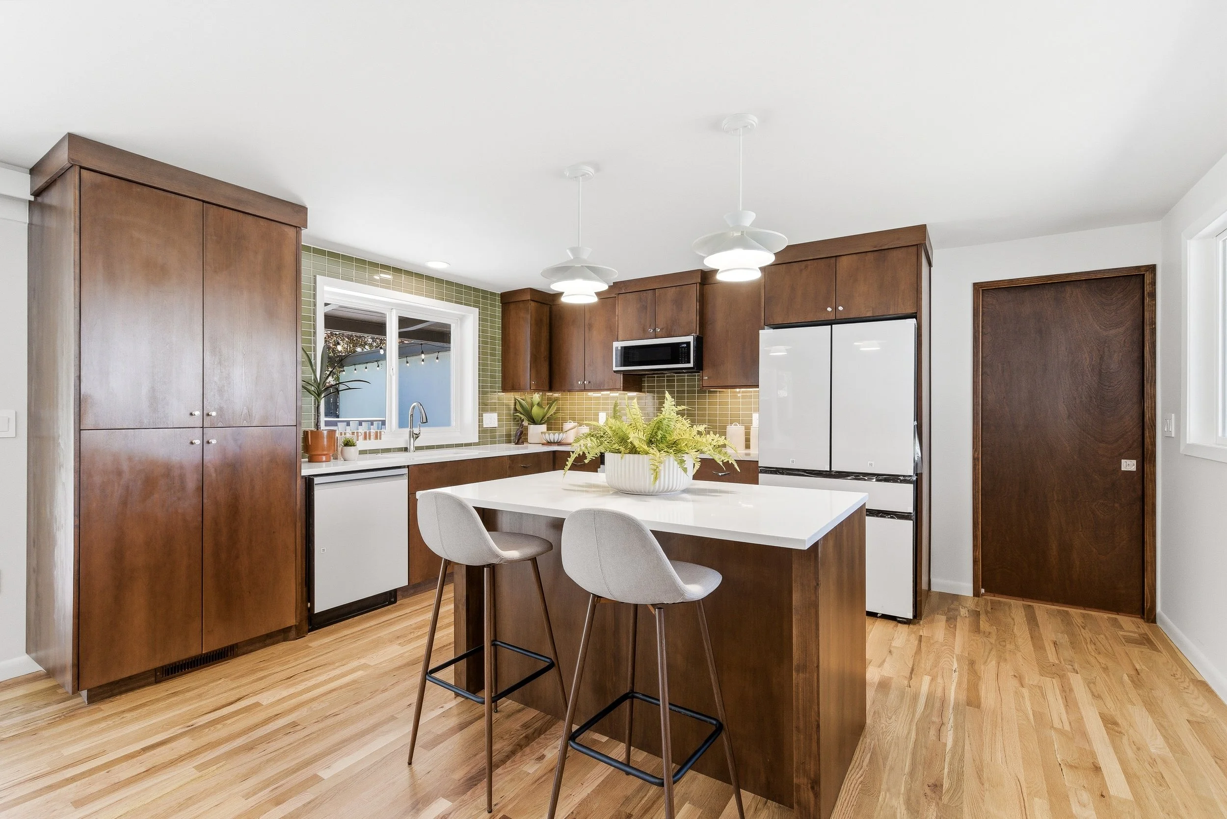 Modern kitchen with white countertops, wood cabinets, a white island with two chairs, green tiled backsplash, window, plants, and pendant ceiling lights.
