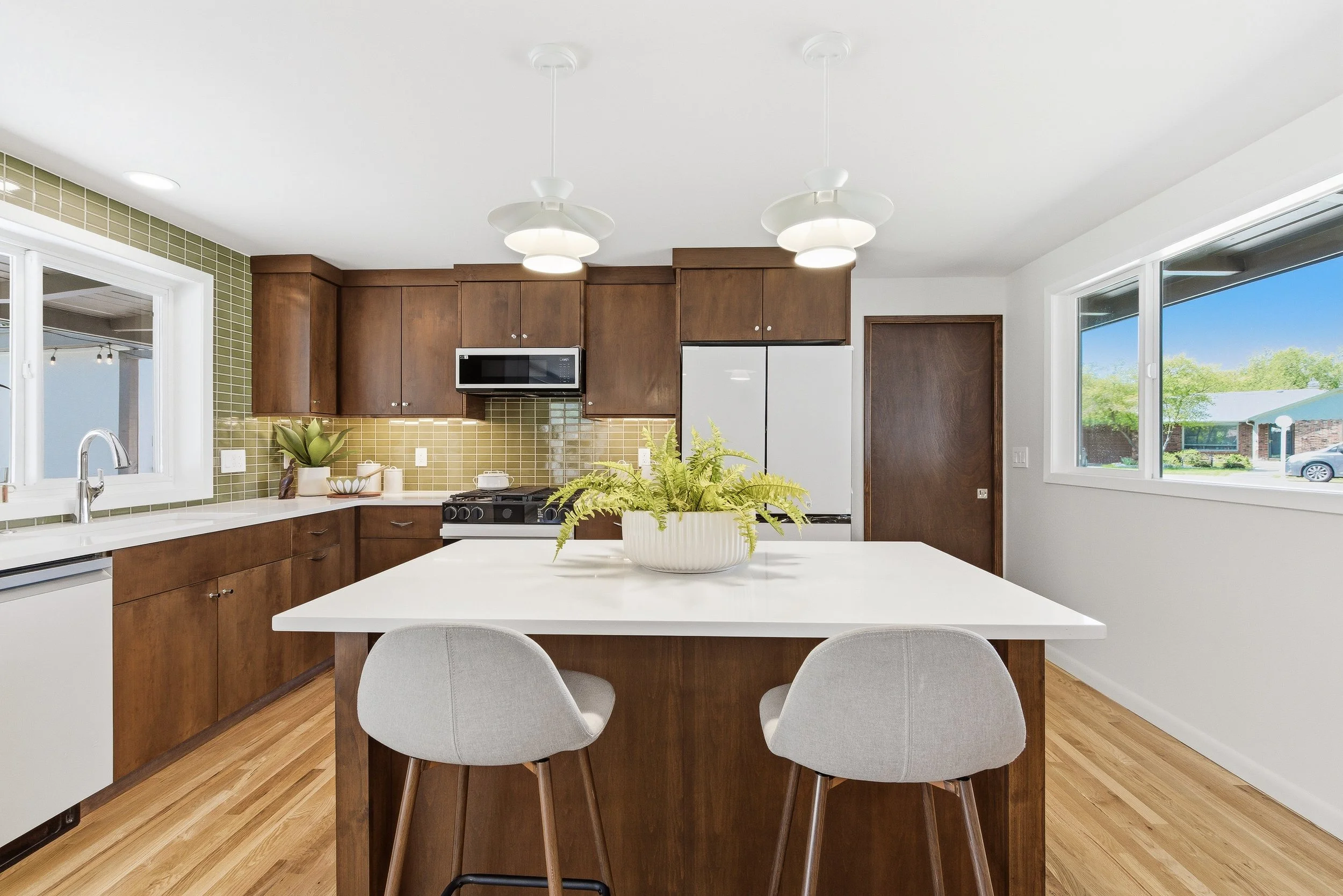 Modern kitchen with wood cabinets, yellow tile backsplash, white countertop, and pendant lighting, dining area with table and chairs, large windows showing a neighborhood outside.