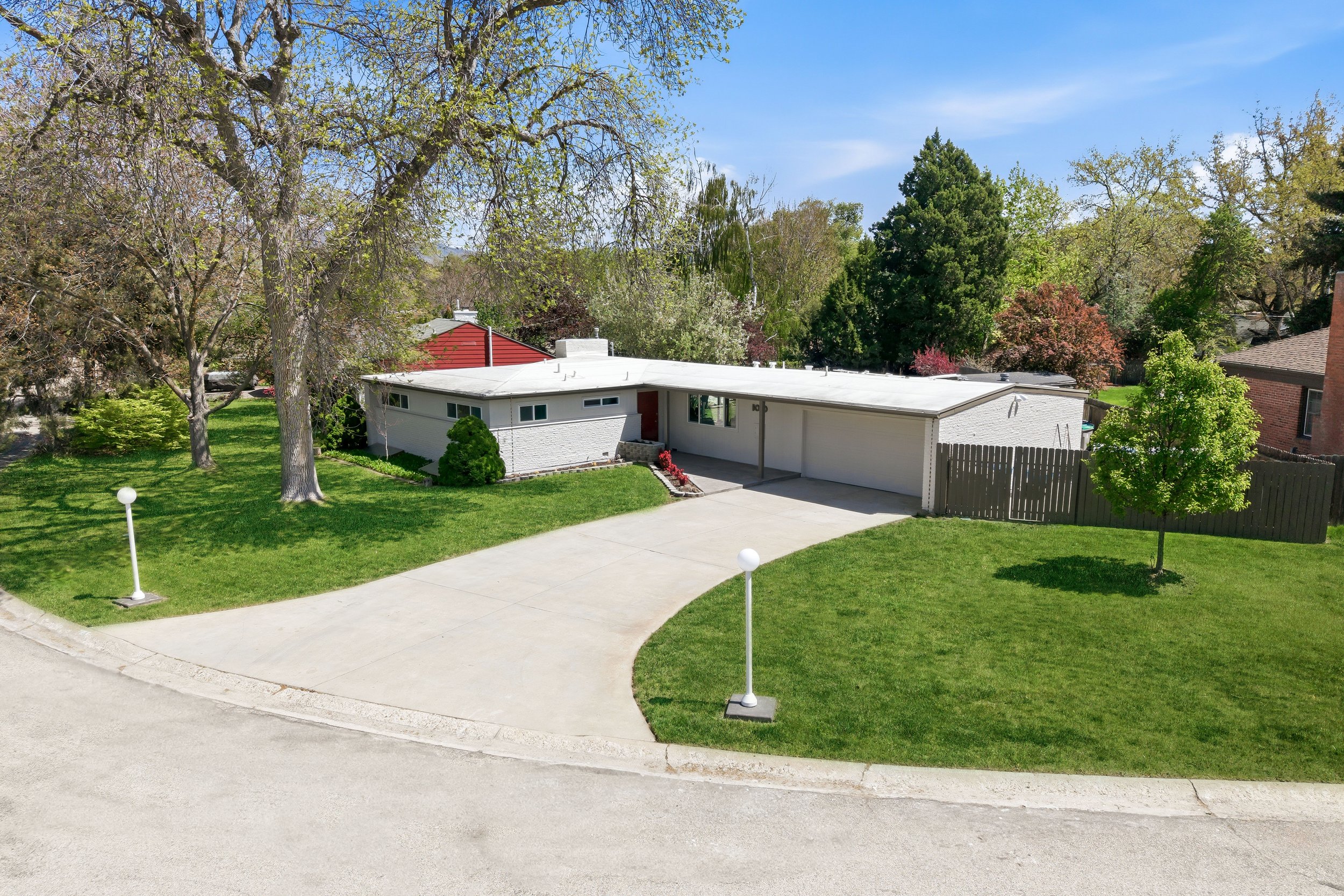 A suburban house with a white exterior, a driveway, and a well-maintained lawn with two small trees. The house has a flat roof and multiple windows. There are trees with green and some reddish leaves, and the sky is clear blue with a few wispy clouds