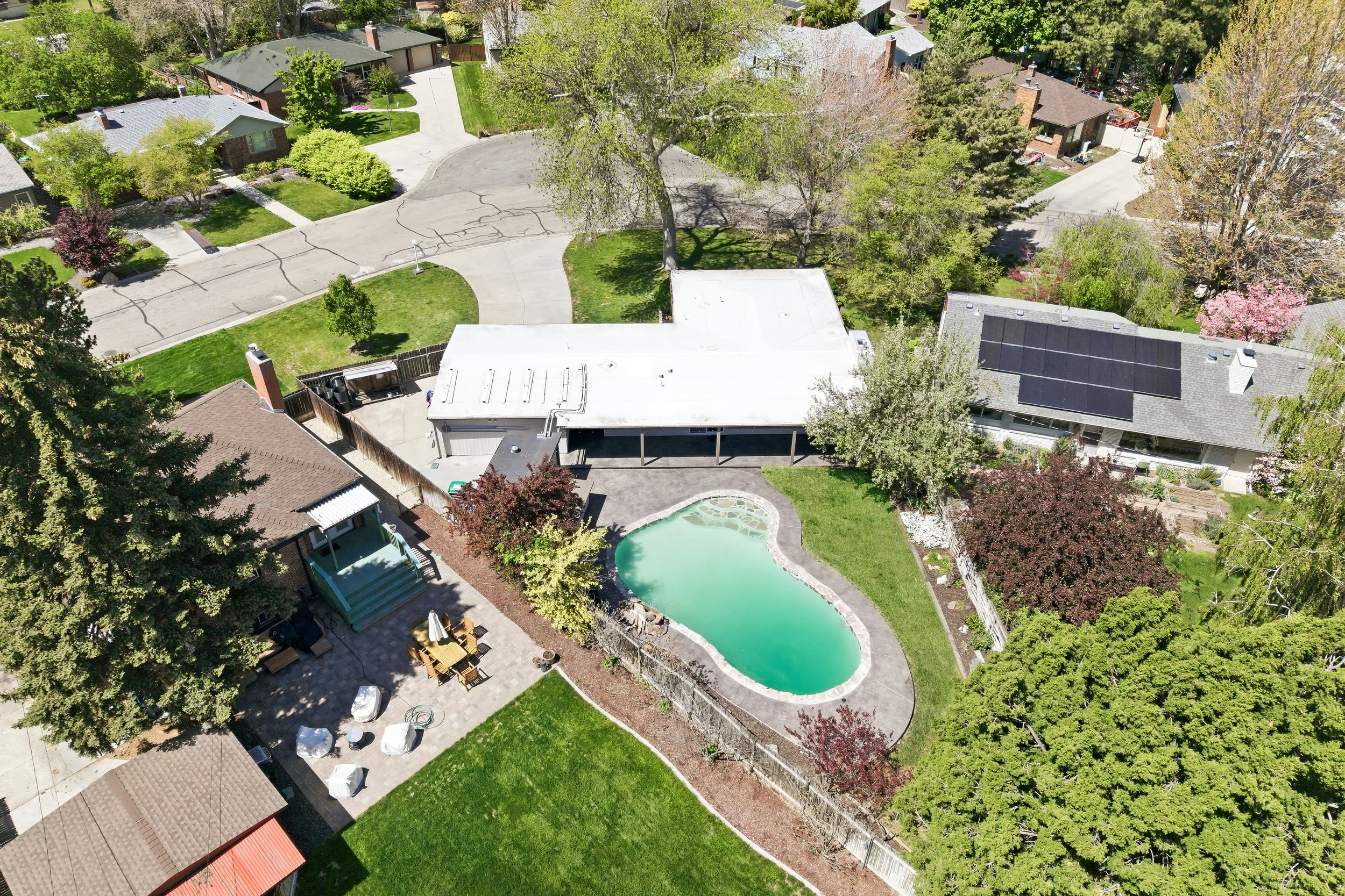 Aerial view of a suburban backyard with a swimming pool, surrounded by trees and landscaped lawn. There is a patio area with outdoor furniture and a small deck with an umbrella.