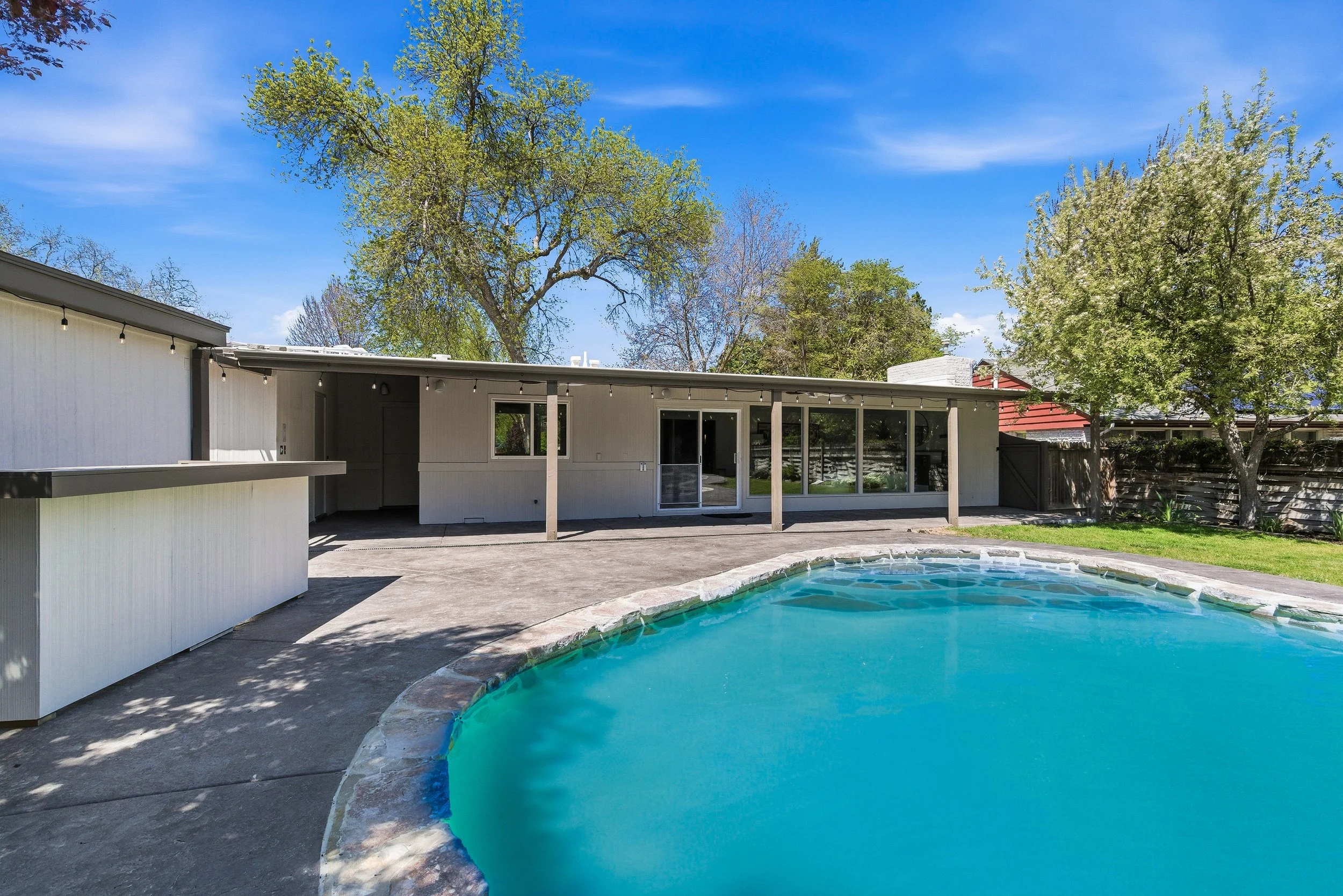 View of a backyard with a small swimming pool, a modern house with large glass doors, a gray fence, green trees, and a clear blue sky.