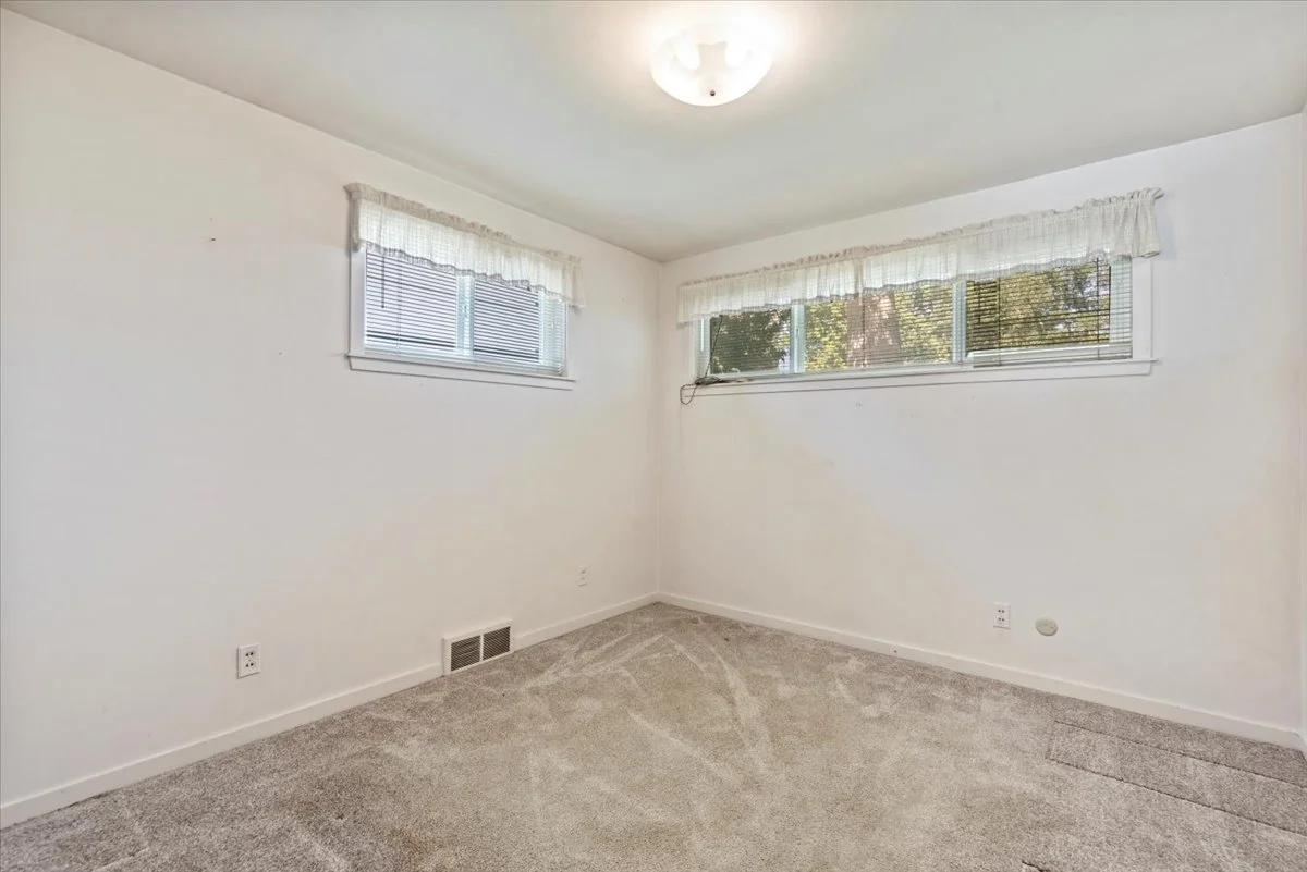 Empty room with white walls, beige carpet, two small windows with blinds and white curtains, and a ceiling light fixture.