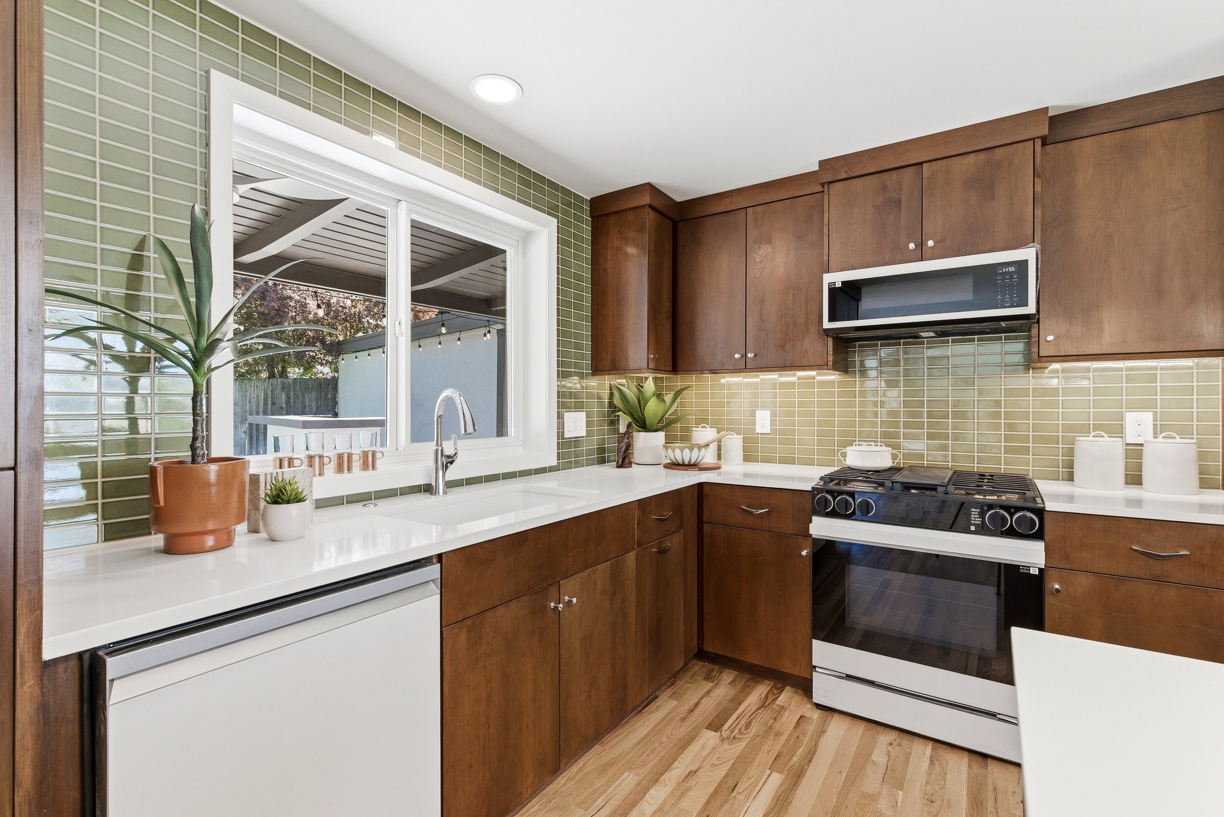 Modern kitchen with brown wooden cabinets, green tile backsplash, white countertops, and stainless steel appliances, viewed through a window showing outdoor space with string lights.
