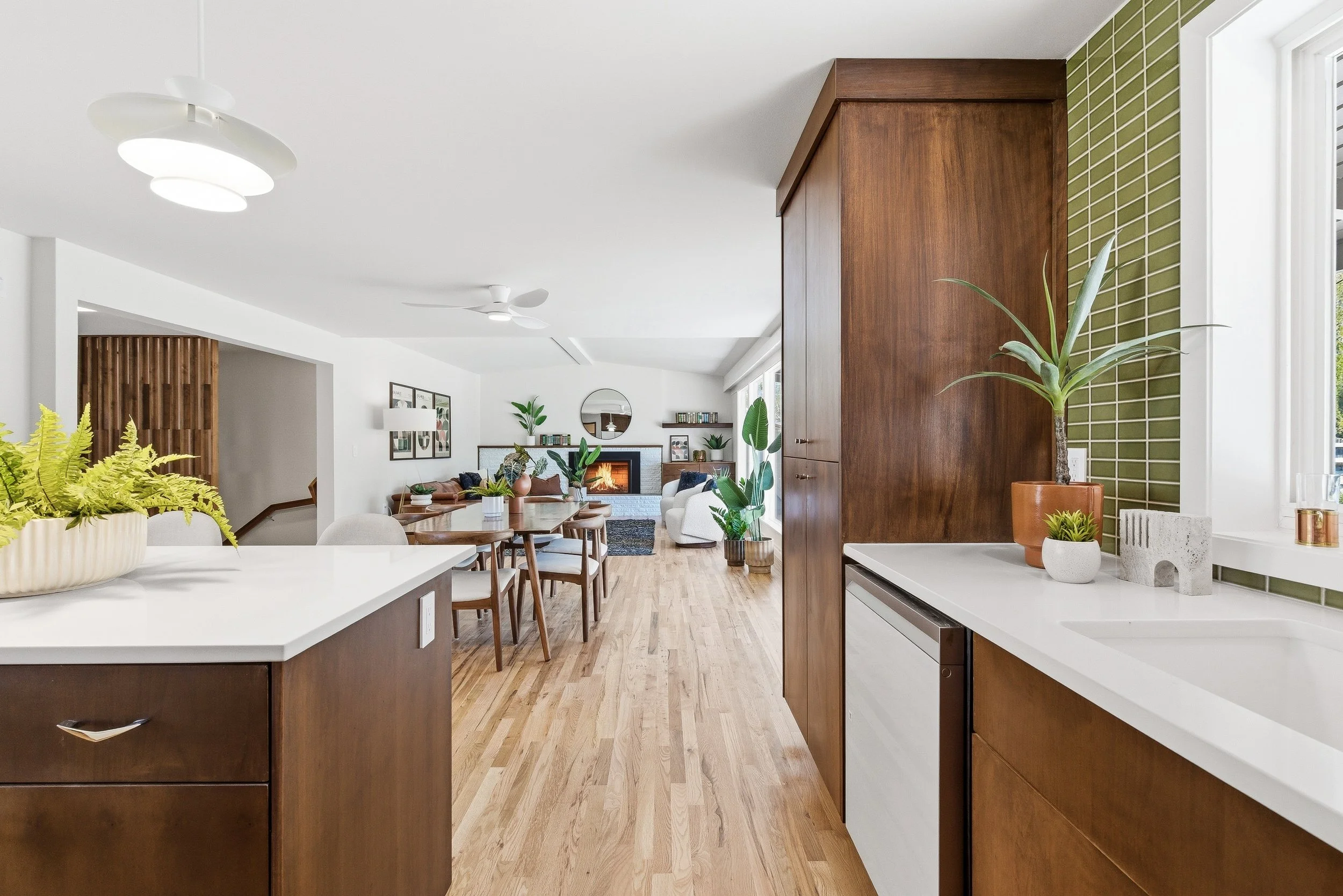 Open-concept living and dining room with wood floors, white walls, modern lighting, green plants, a fireplace, and large windows.