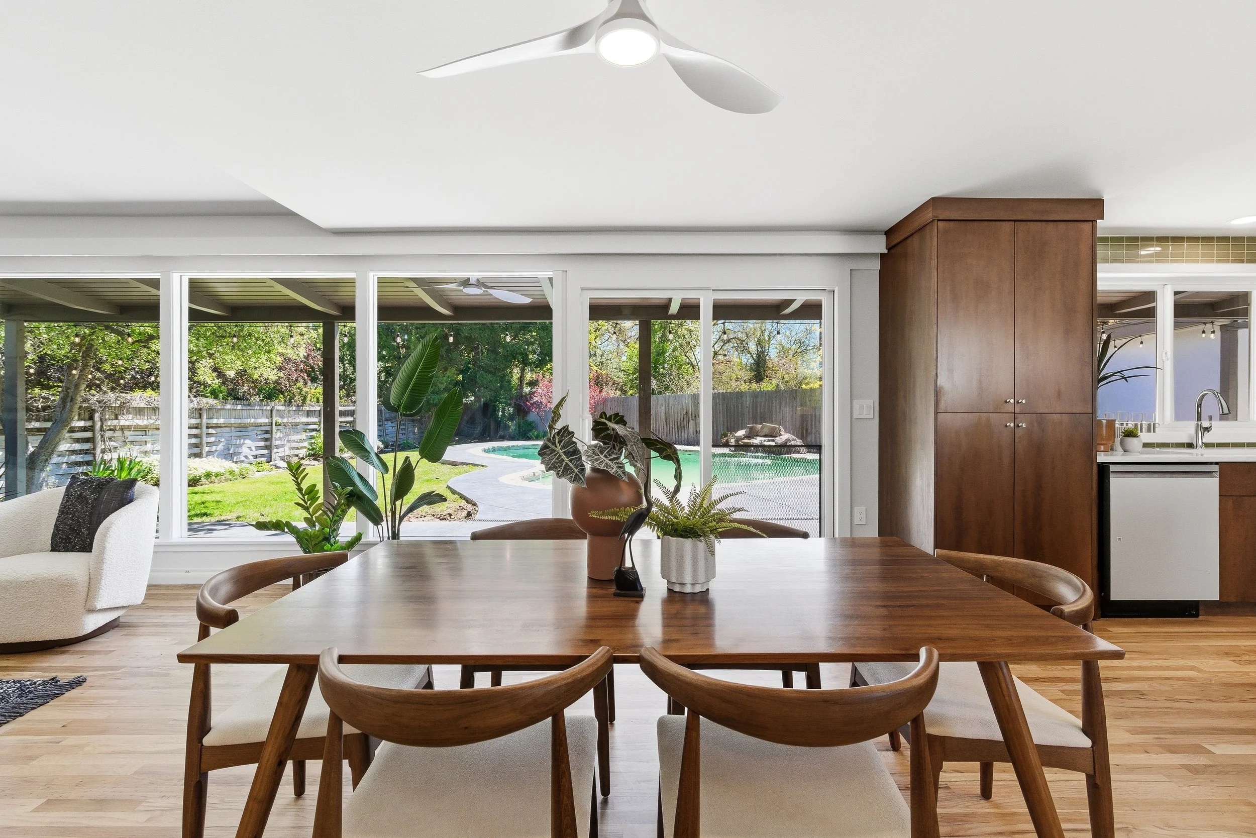Interior view of a living room and kitchen area with a wooden dining table, potted plants, large glass doors revealing a backyard with a pool, backyard fence, trees, and outdoor seating, a ceiling fan, and a white ceiling.