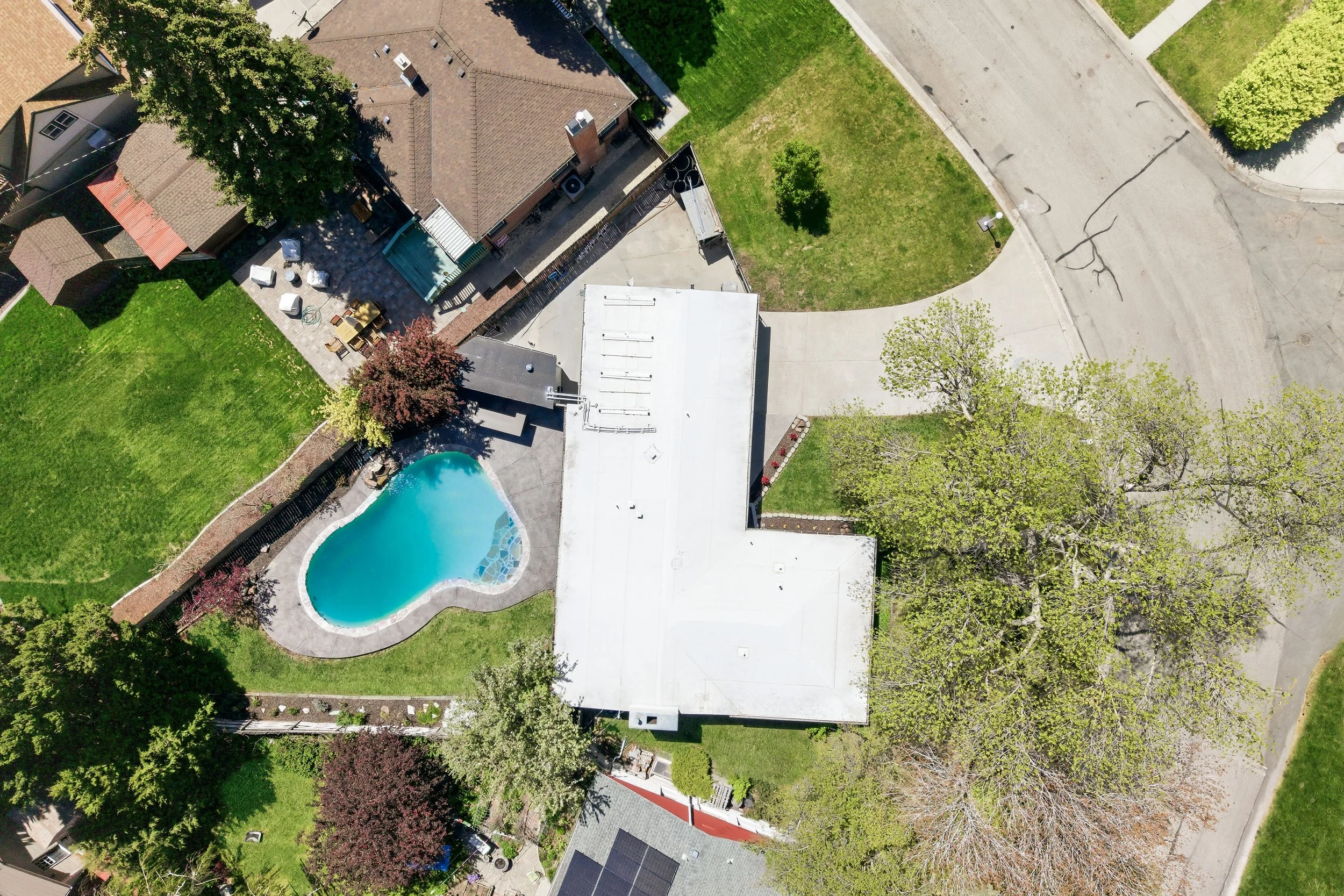 An aerial view of a suburban backyard with a kidney-shaped swimming pool, surrounded by a concrete patio. The backyard features a green lawn with trees, a garden area, and outdoor furniture including a table with chairs. The house has a brown roof wi
